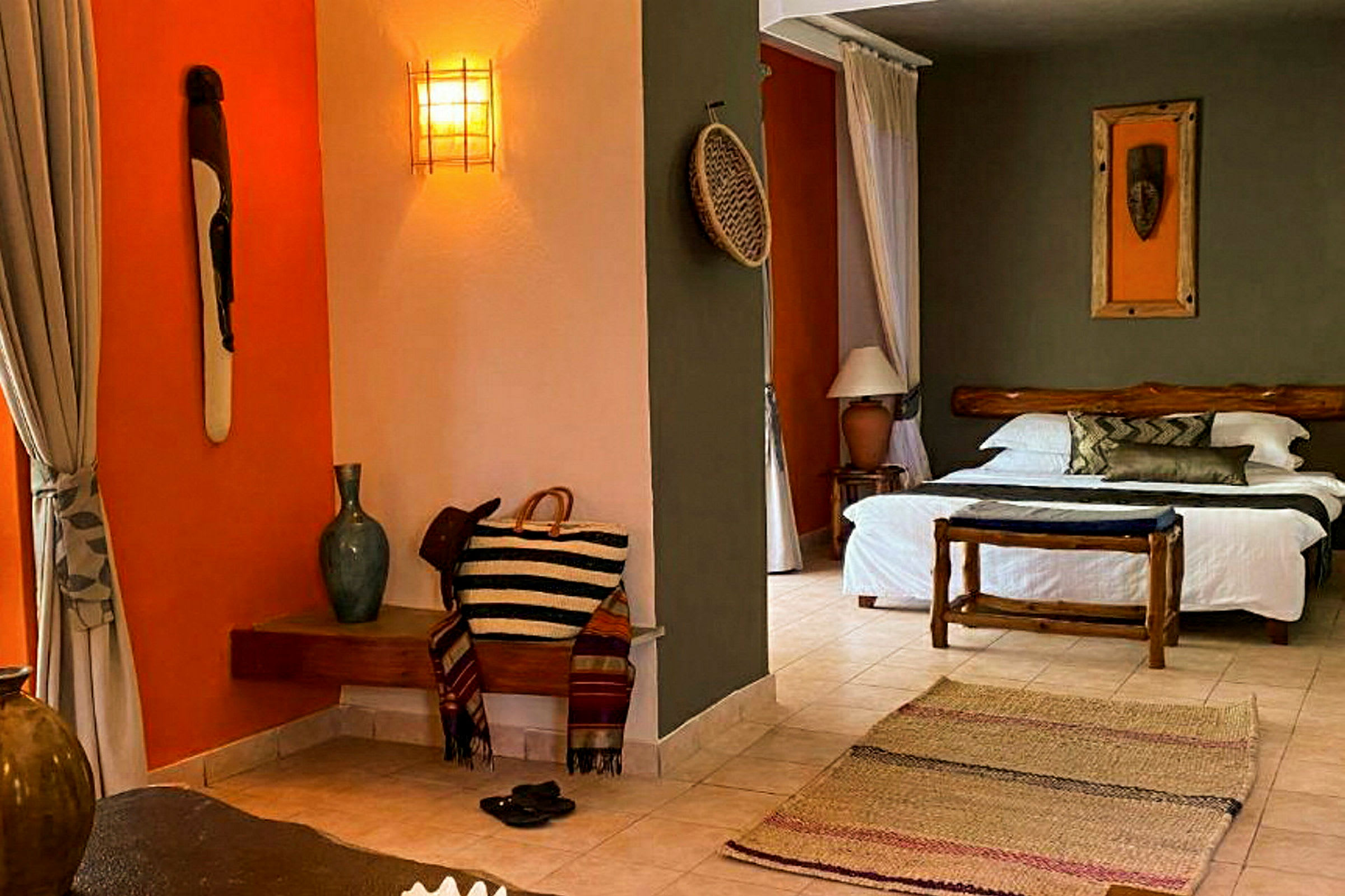 En hotellrumsinteriör med en dubbelsäng, en träbänk, en tillhörande dekorativ matta och accentdetaljer som maskkonst och orangefärgade väggar. (AI-genererad text)