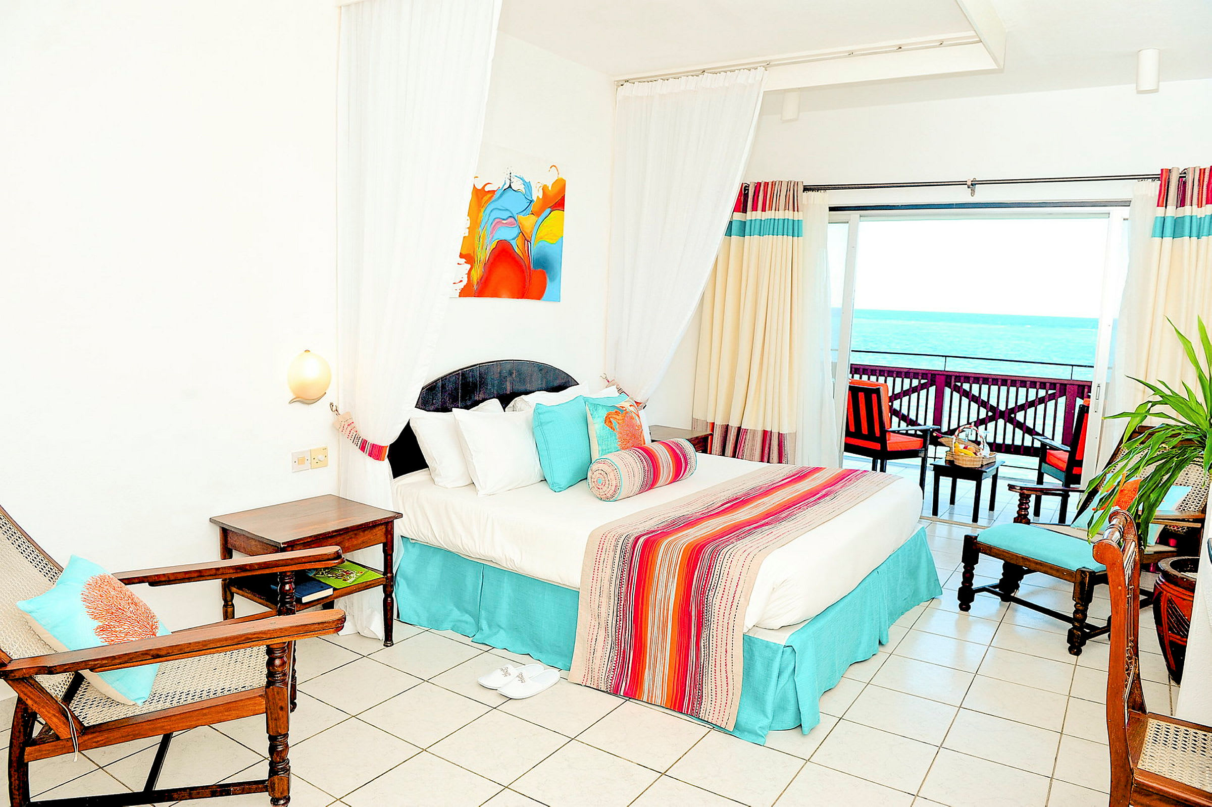 Ett hotellrum med en stor säng, färgglad överkast och dekorativa kuddar, ett rustikt träbord med stolar och en balkong med utsikt över havet. (AI-genererad text)