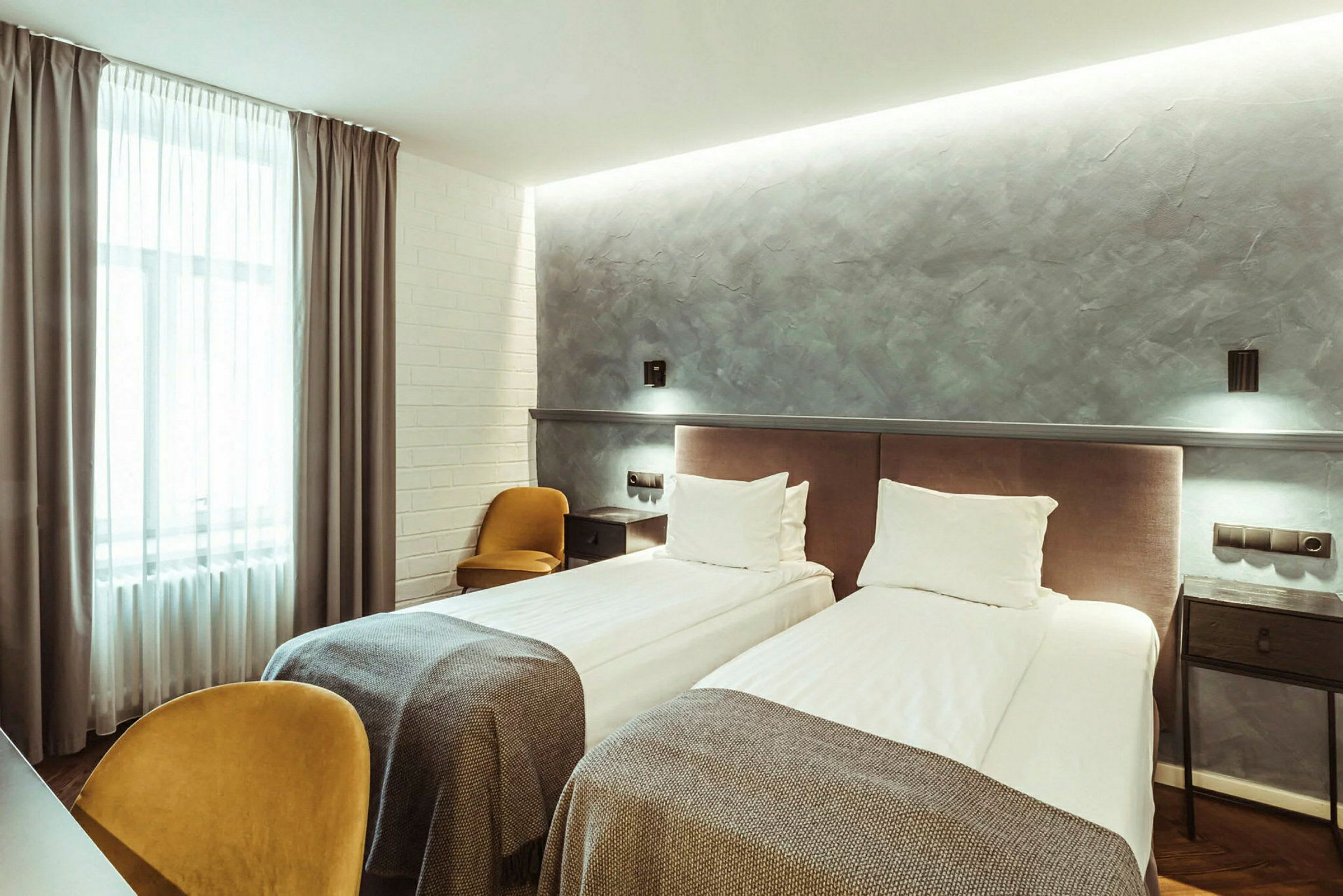 Ett hotellrum med två enkelsängar, en grå vägg och en stor fönstergardin. (AI-genererad text)