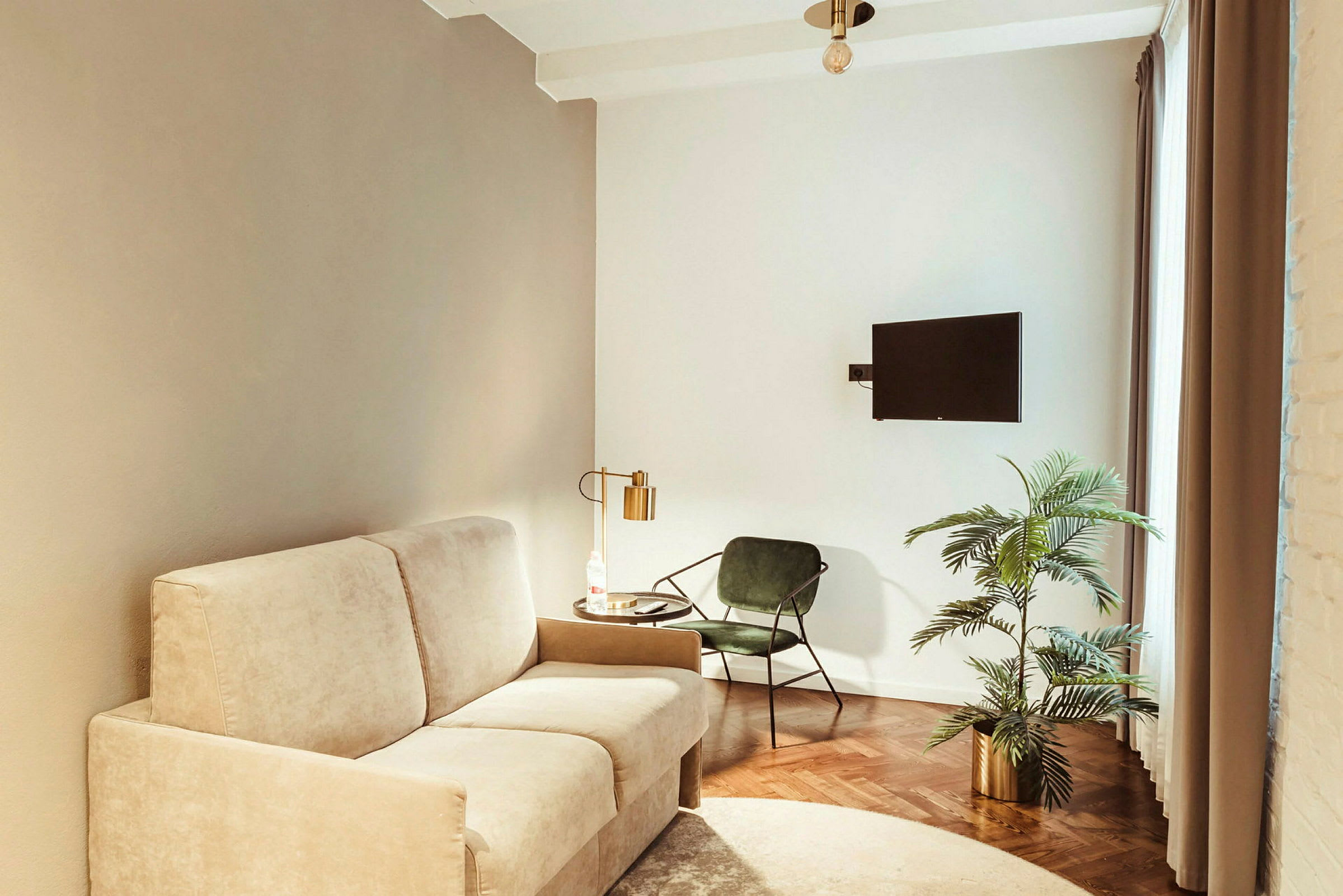 Ett vardagsrum med beige soffa, grön stol, bord med lampa, växt i kruka och väggmonterad TV. (AI-genererad text)