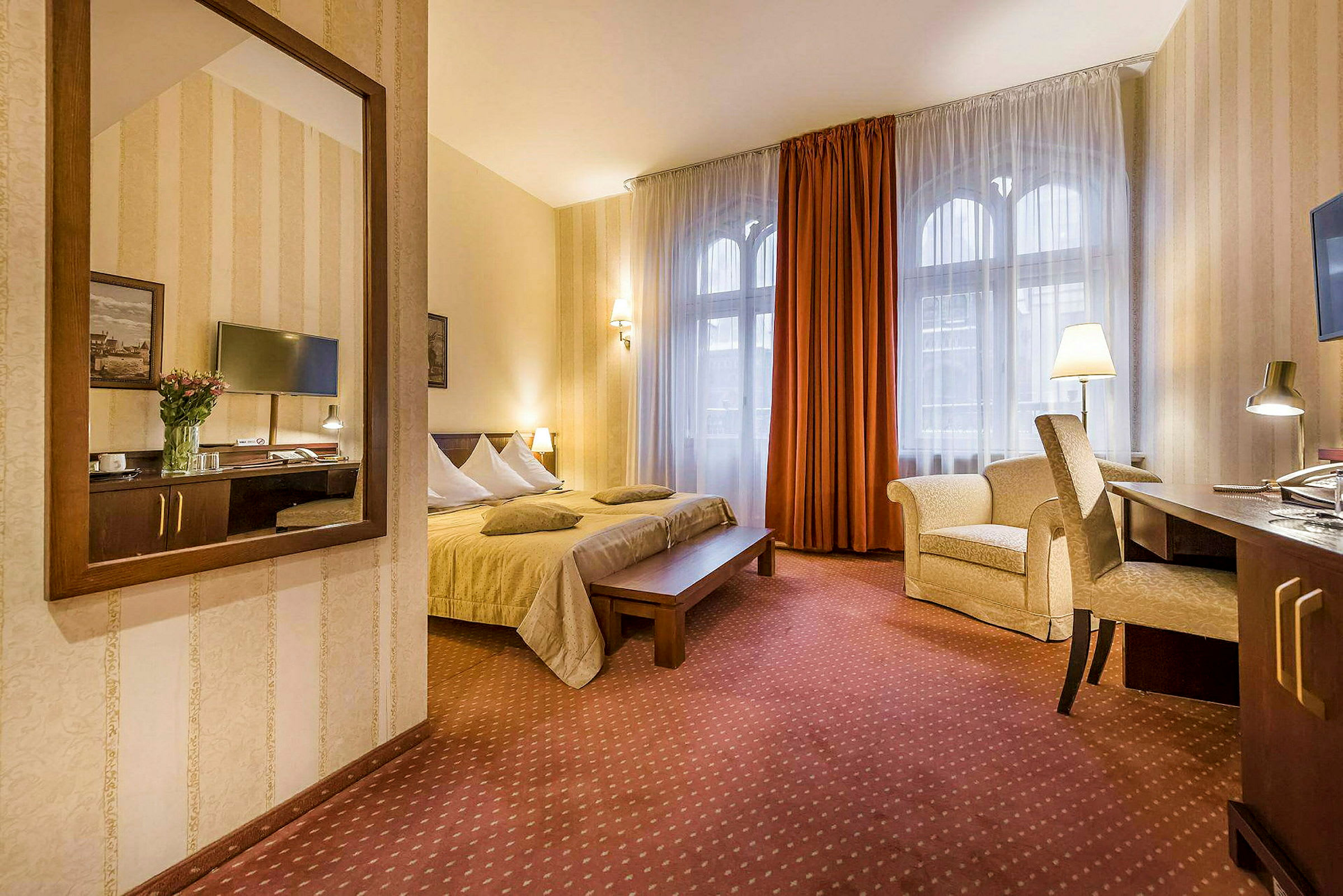 Ett hotellrum med en dubbelsäng, skrivbord och stol, och en fåtölj. Fönster med gardiner och en väggspegel. (AI-genererad text)