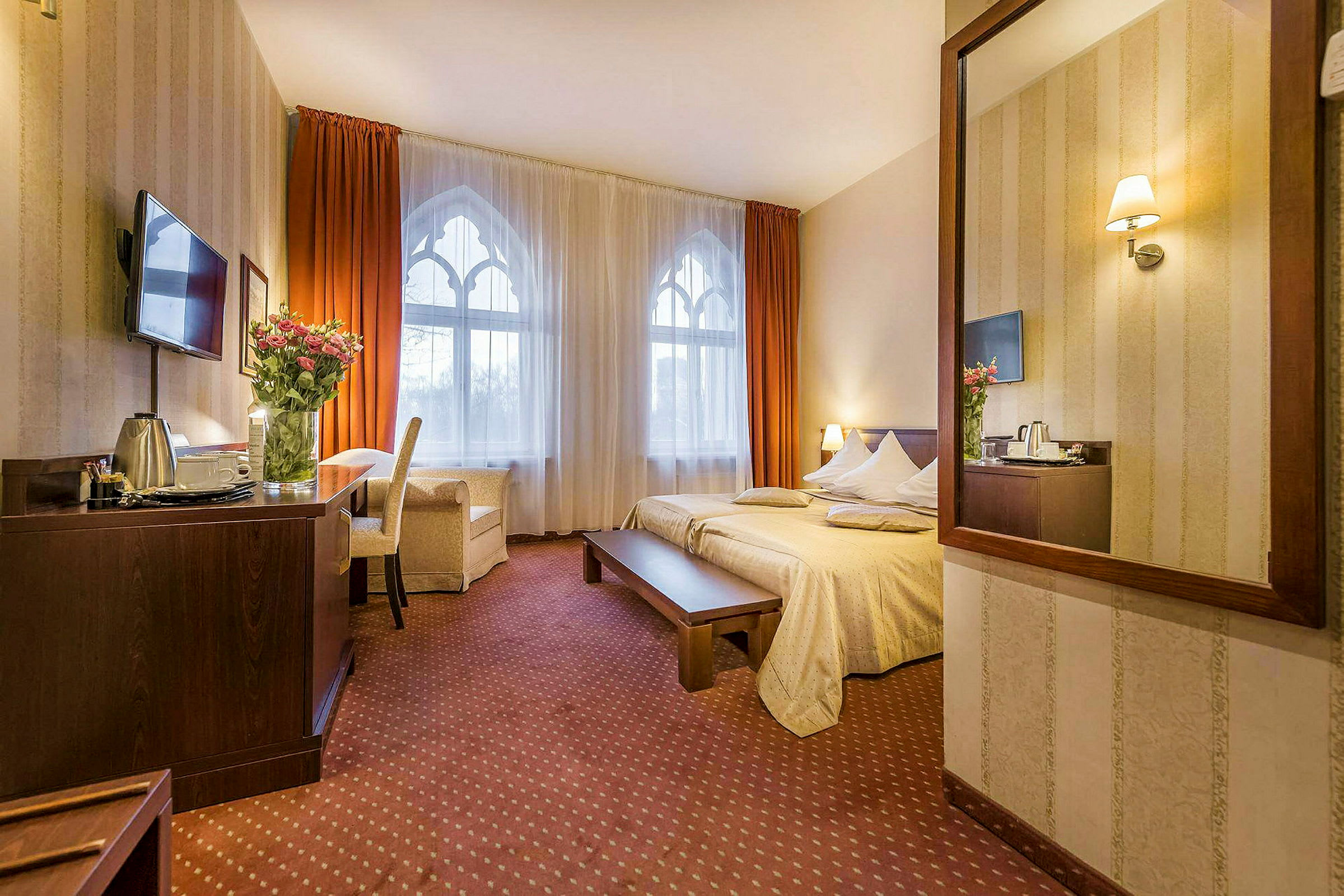 Ett hotellrum med en stor säng, ett bord och en stol. Fönstret har vita gardiner med orangea draperier. (AI-genererad text)