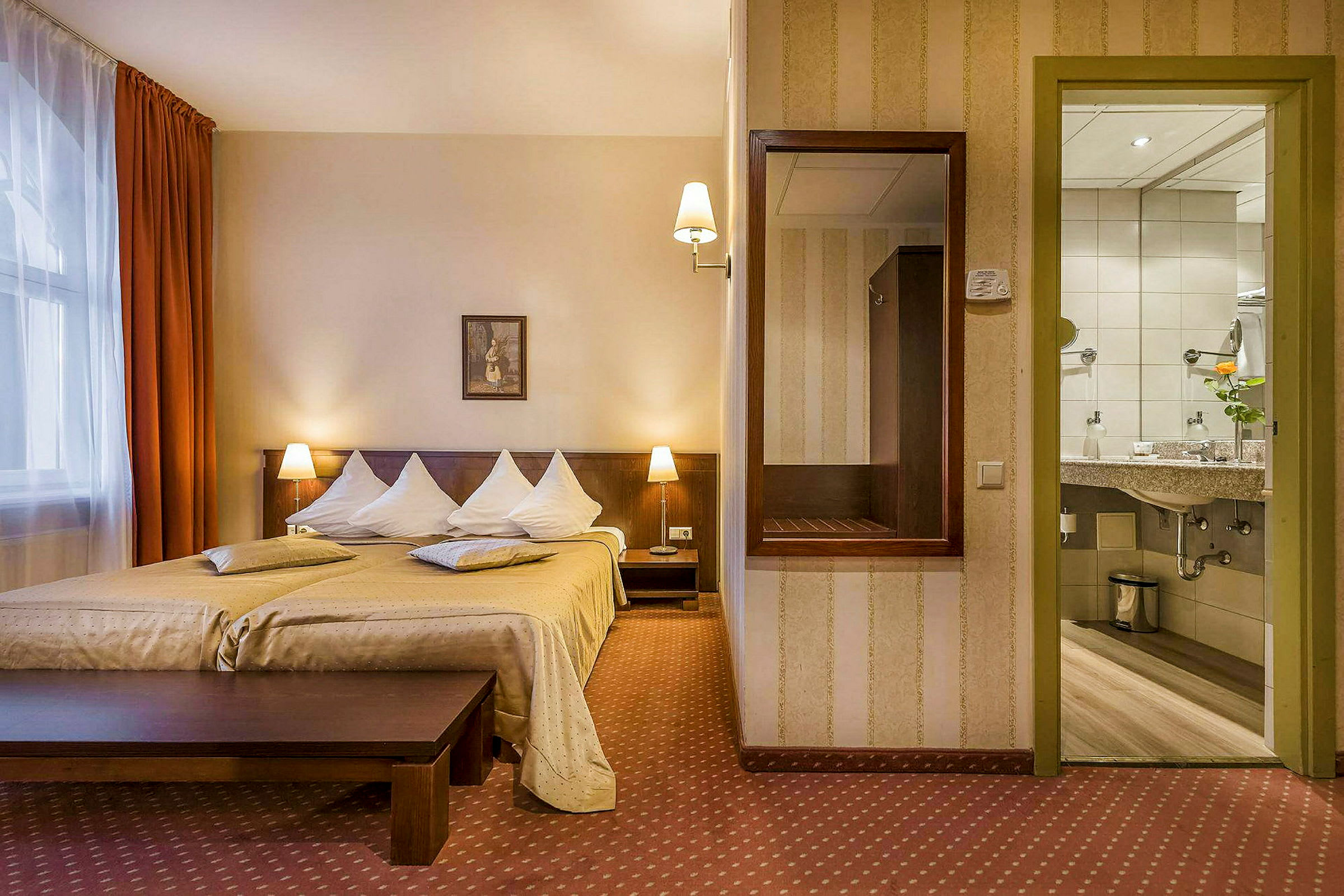 Ett hotellrum med en dubbelsäng, två nattduksbord med lampor, en spegel på väggen och en vy mot badrummet. (AI-genererad text)