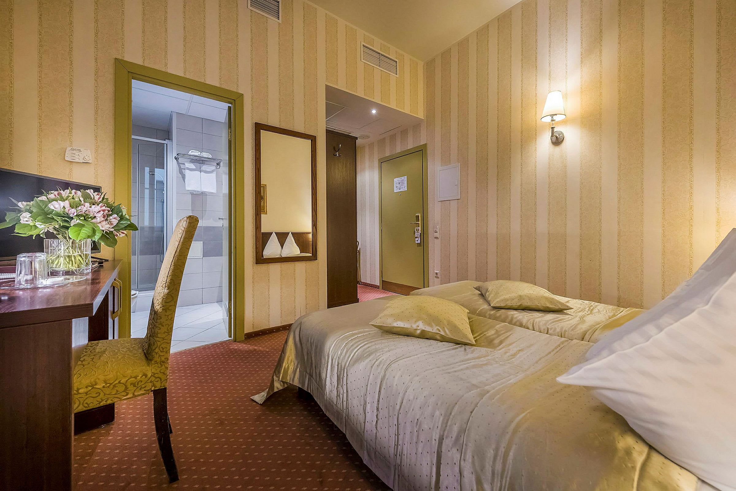 Ett hotellrum med en bäddad säng, ett skrivbord med en blomvas och en stol samt dörrar till badrum och hall. (AI-genererad text)