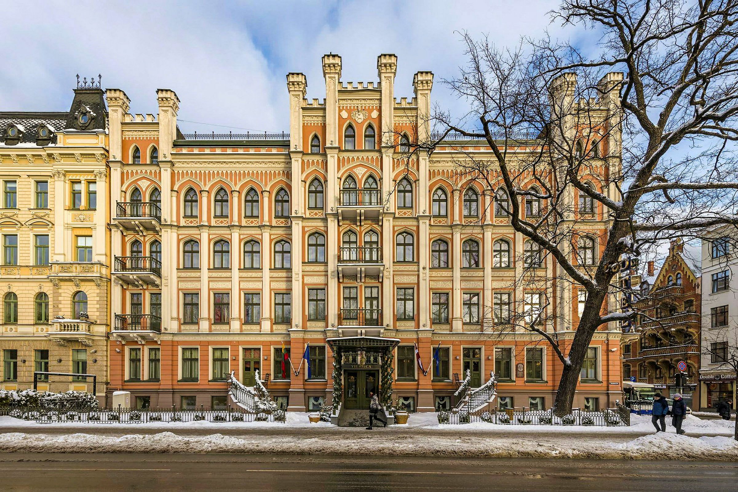 En fasad av en ståtlig byggnad med gotisk arkitektur och flera fönster, sett under vintertid med snö på marken. (AI-genererad text)