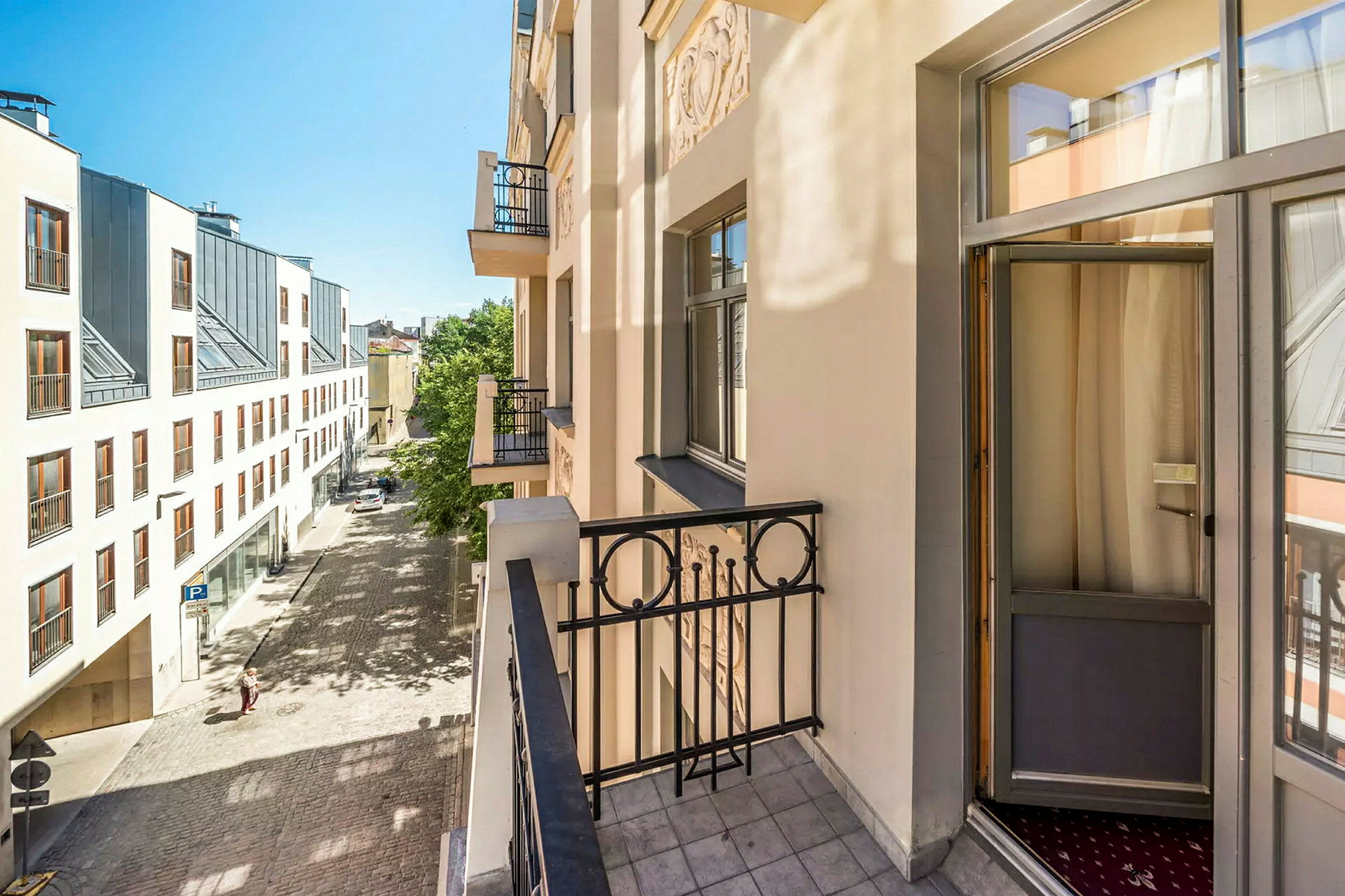Bilden visar en balkong på en ljus byggnad med utsikt över en stenlagd gata och ett modernt hus med stora fönster på andra sidan gatan. (AI-genererad text)