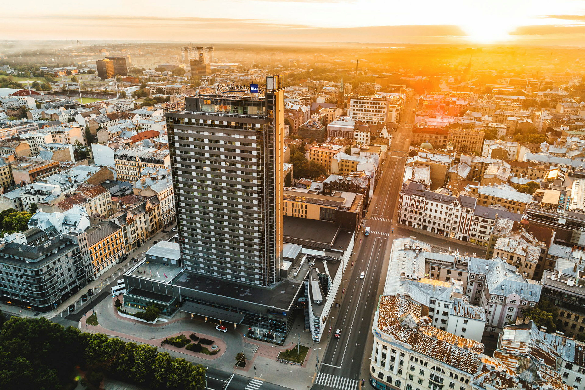 Flygfoto av en stad med en hög byggnad i framgrunden och andra byggnader omkring, samtidigt som solen går ner vid horisonten. (AI-genererad text)