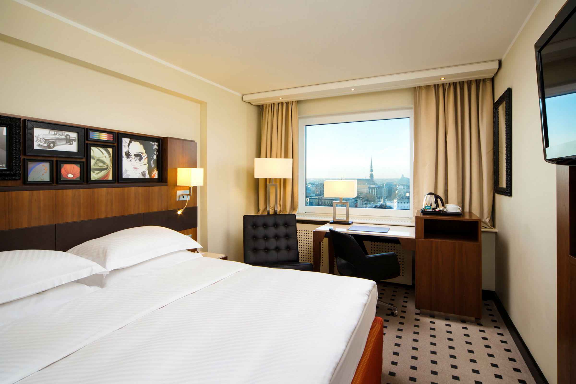 Hotellrum med en stor säng framför ett skrivbord med utsikt över en stad genom ett fönster. (AI-genererad text)
