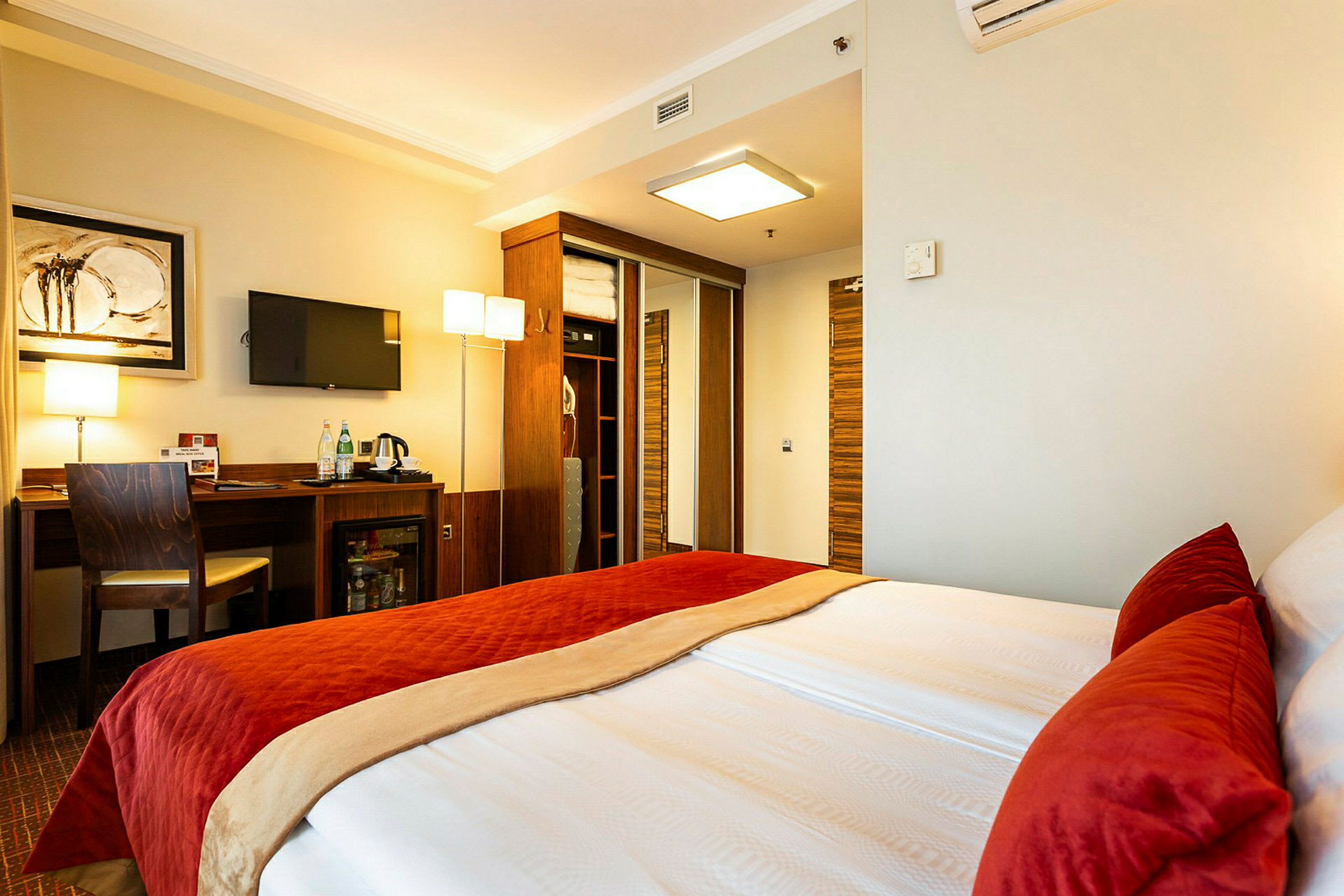 Ett hotellrum med en dubbelsäng, inrett med röda och beige sängkläder. Rummet har ett skrivbord med en stol, en väggmonterad TV, och en garderob med spegelbeklädda dörrar. (AI-genererad text)