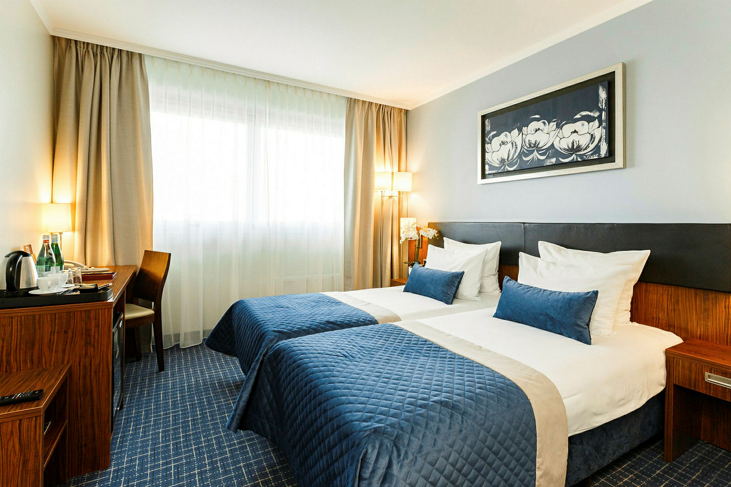 Ett hotellrum med två enkelsängar, blå överkast och huvudgavlar. Ett skrivbord med stol står bredvid, samt en vattenkokare och flaskor. (AI-genererad text)