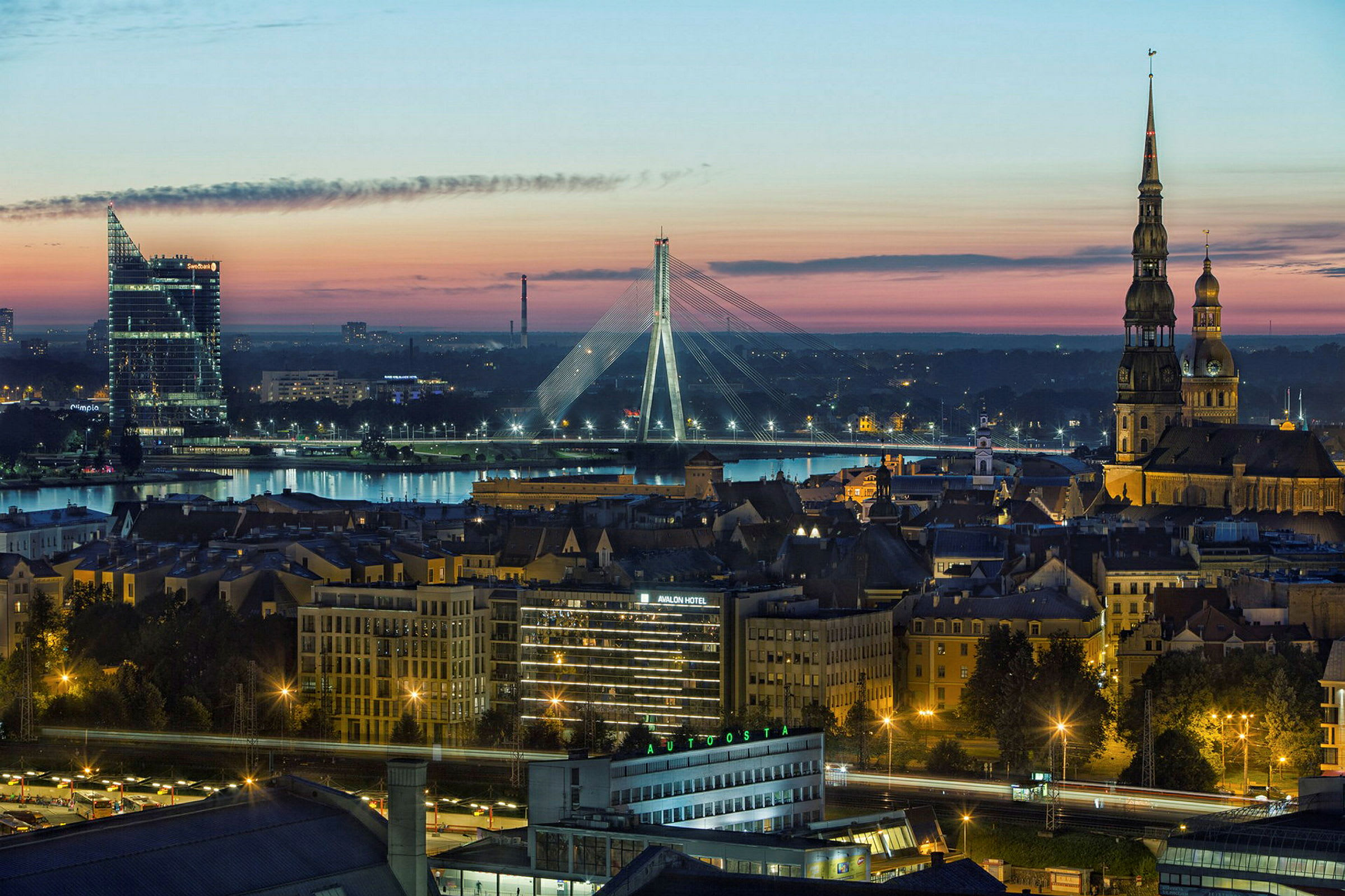 Panoramabild av en stad i skymningen, med en flod, upplysta byggnader och en bro i bakgrunden. (AI-genererad text)