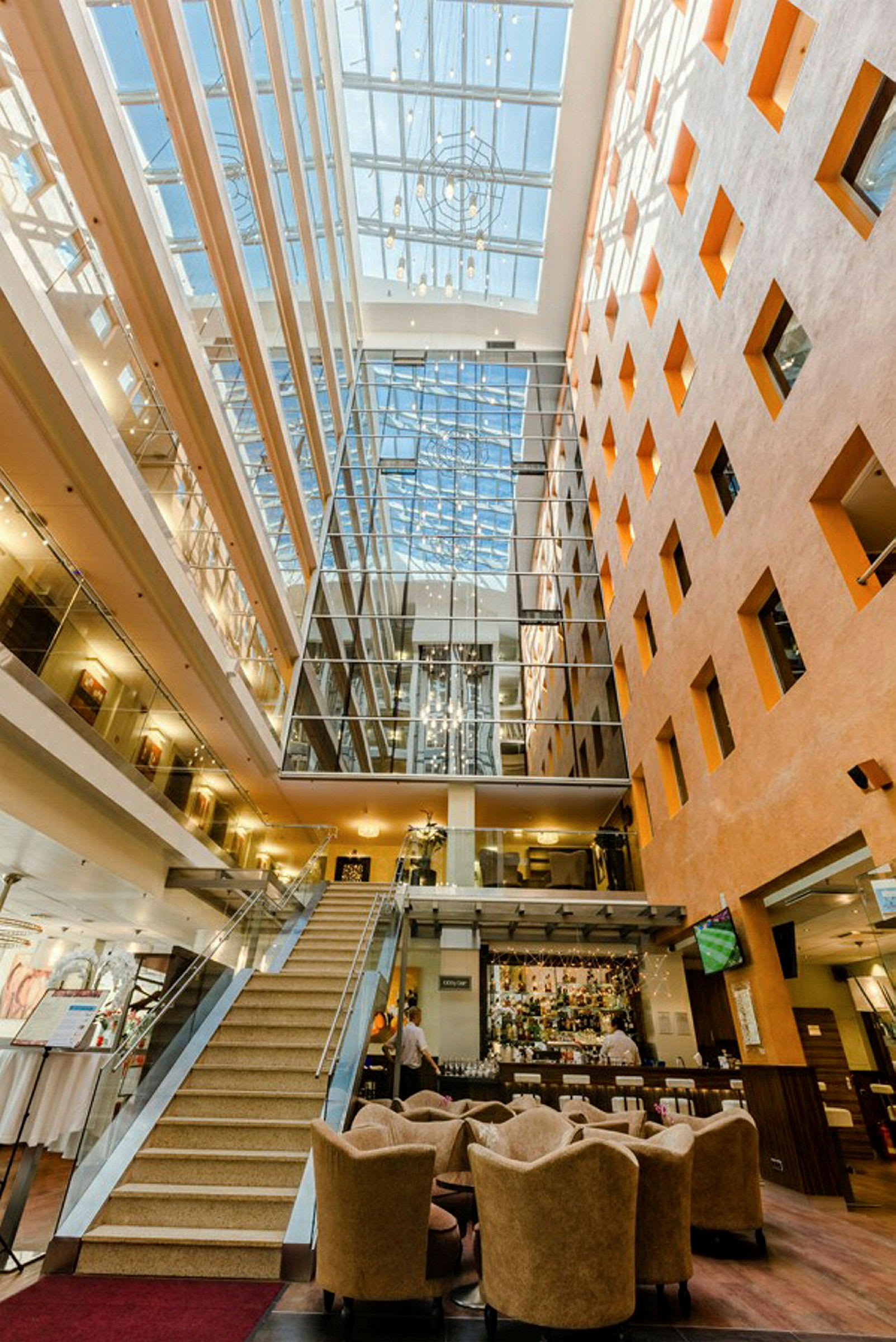 Bild av en ljus hotellfoajé med en trappa, sittgrupper och en reception dekorativt belyst av naturligt ljus från ett takfönster. (AI-genererad text)