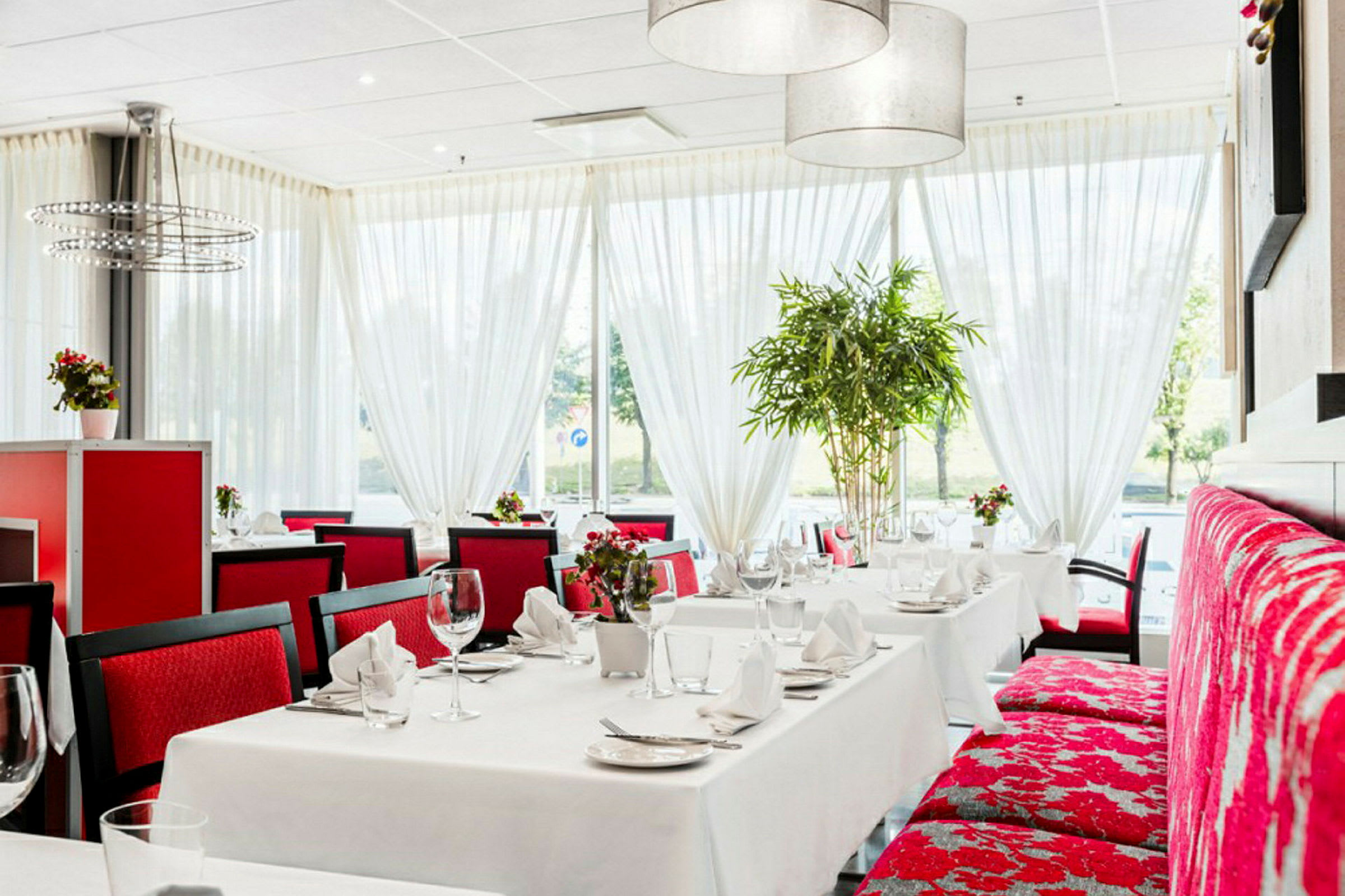 En restaurang med vita dukar och röda stolar, dekorerad med stora fönster och gröna växter. (AI-genererad text)
