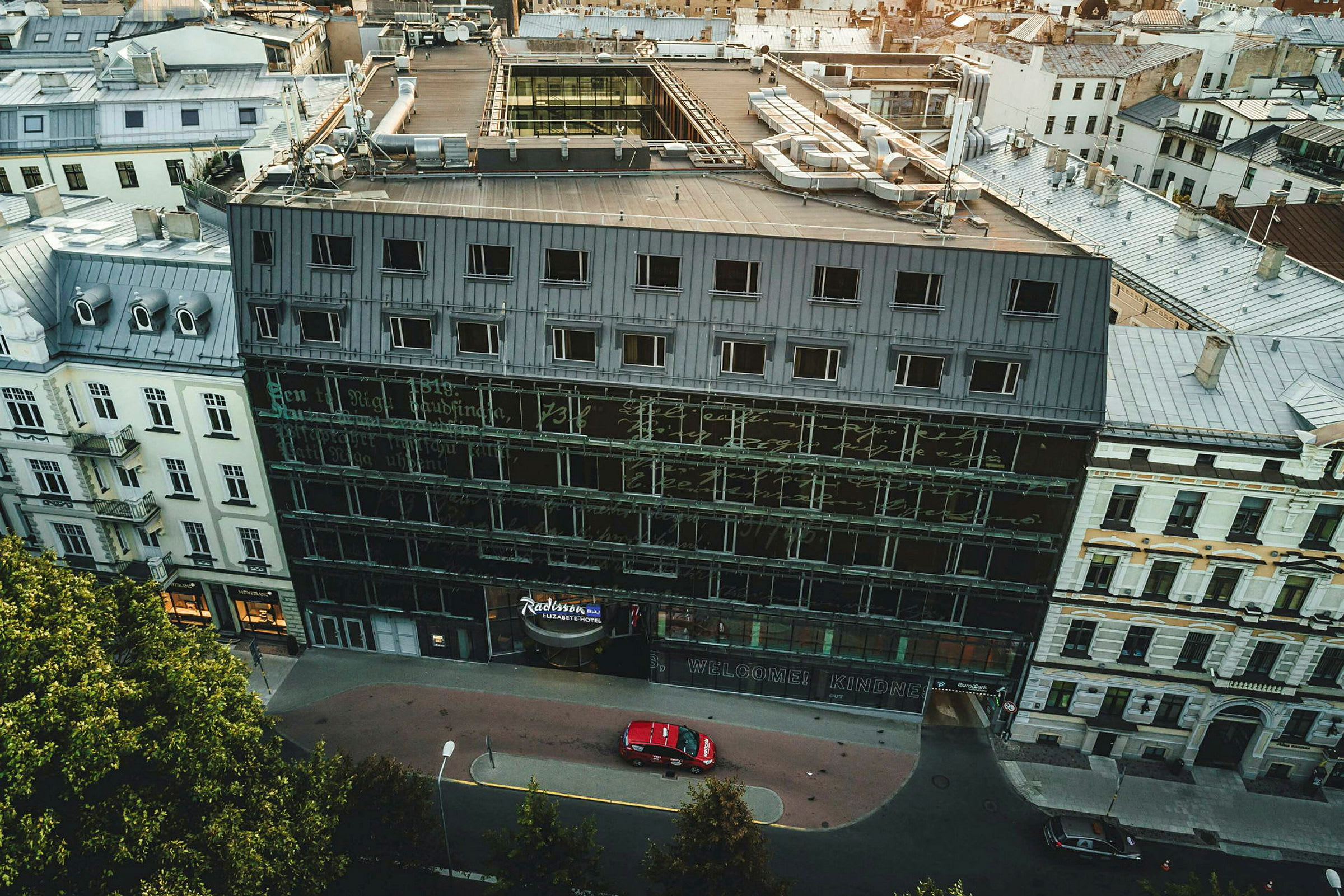 Flygfoto av en byggnad med glasfasad och flera våningar omgiven av andra byggnader. (AI-genererad text)