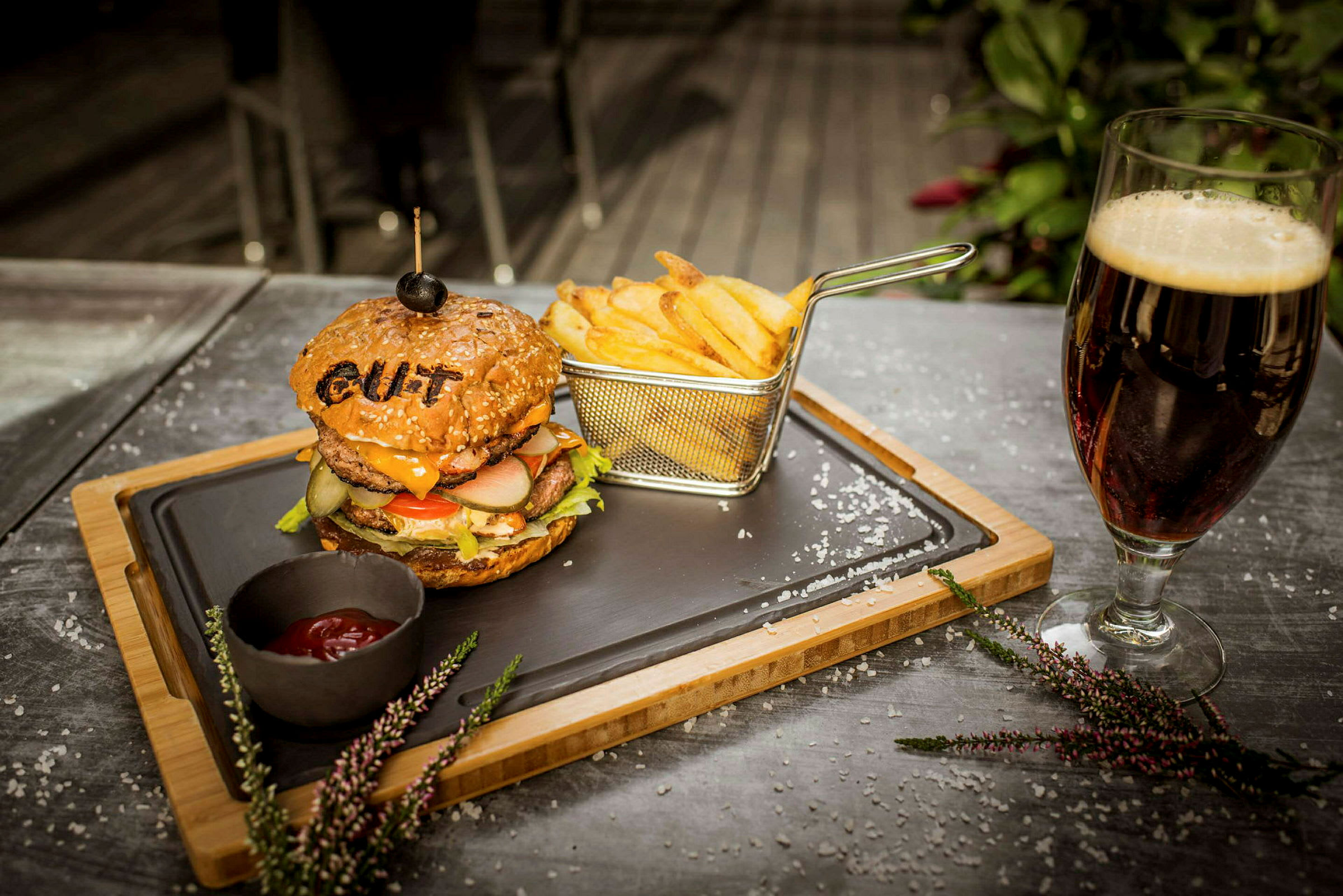 En tallrik med en hamburgare och pommes frites samt en skål med ketchup, placerad på ett bord bredvid ett glas mörk dryck. (AI-genererad text)