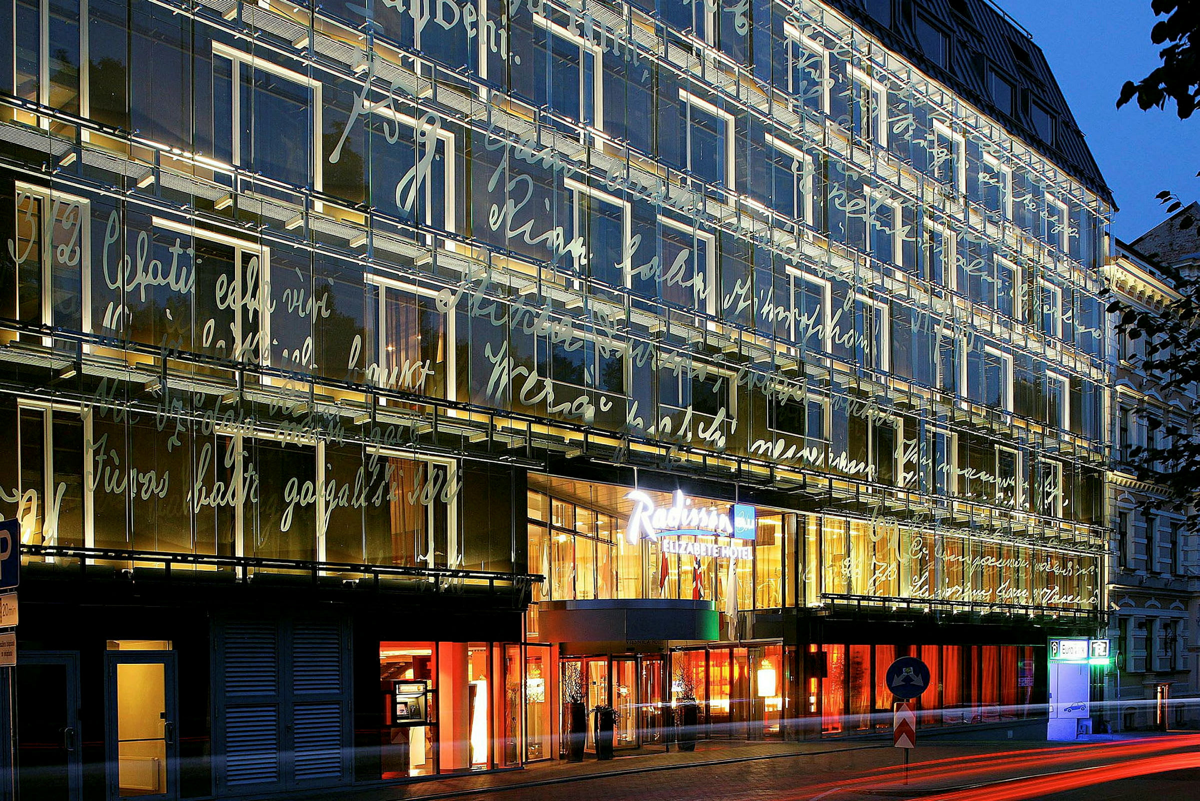 En modern byggnad med glasfasad upplyst på kvällen, dekorerad med skrivstilar. Ingången till en hotellkedja syns tydligt. (AI-genererad text)