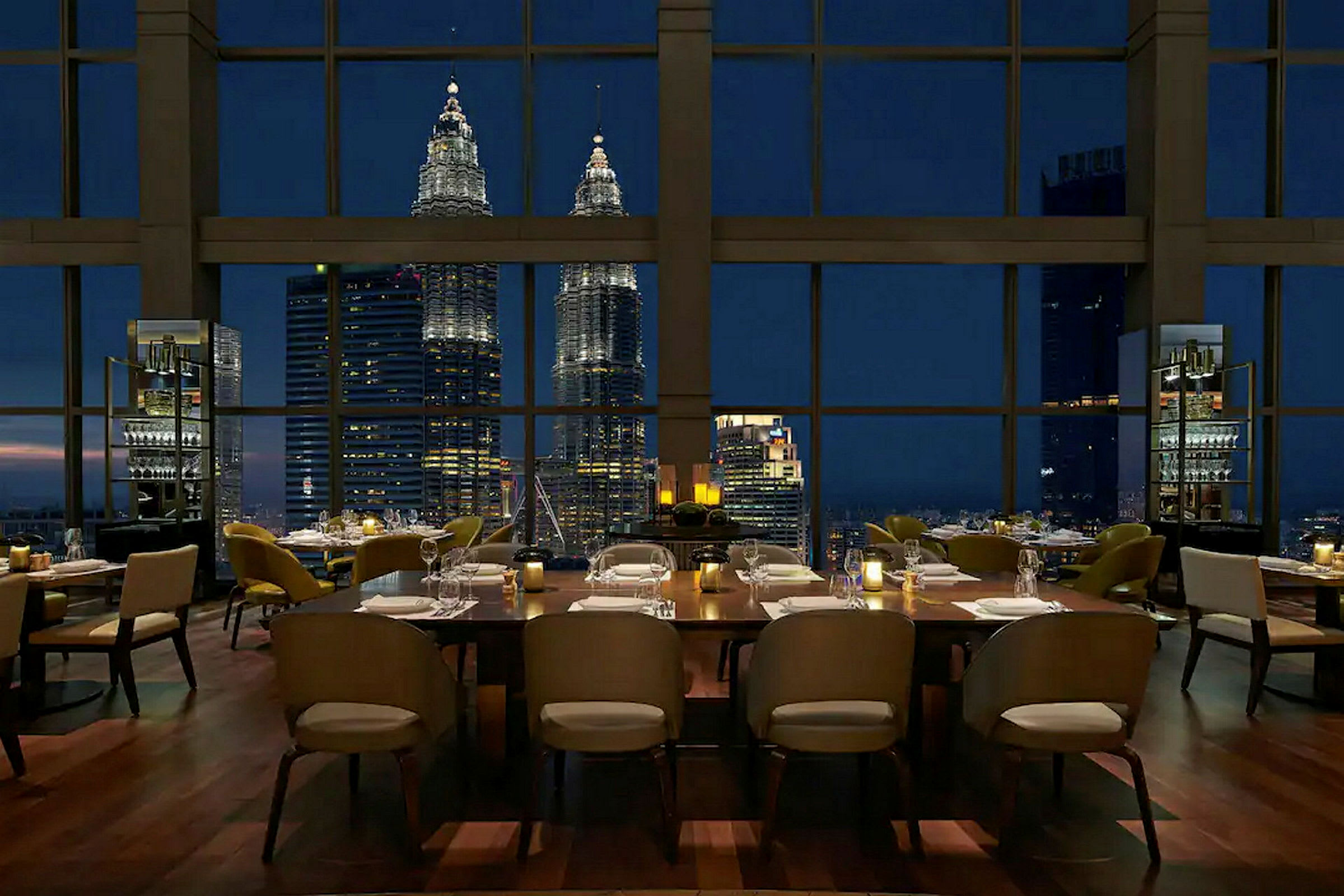 En restaurang med uppdukade bord som har utsikt mot Petronas Twin Towers genom stora fönster på kvällen. (AI-genererad text)