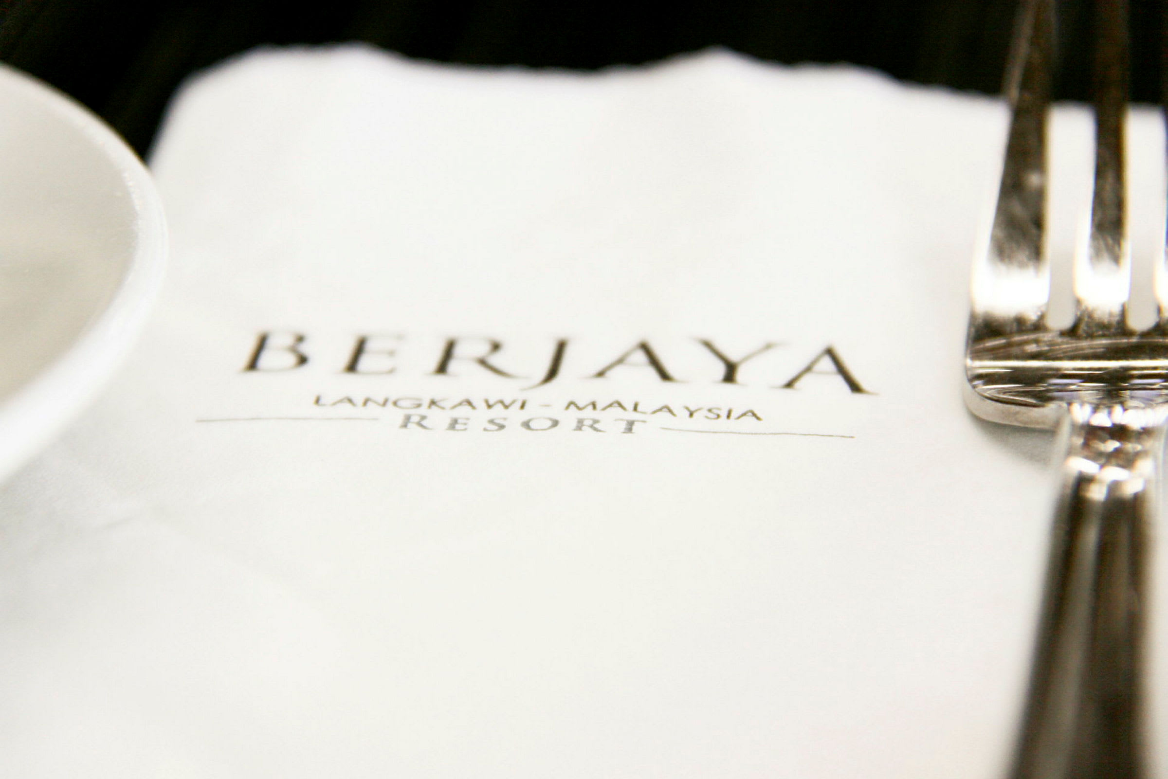 En närbild av en servett med texten "BERJAYA Langkawi - Malaysia Resort" samt en gaffel och del av ett fat. (AI-genererad text)
