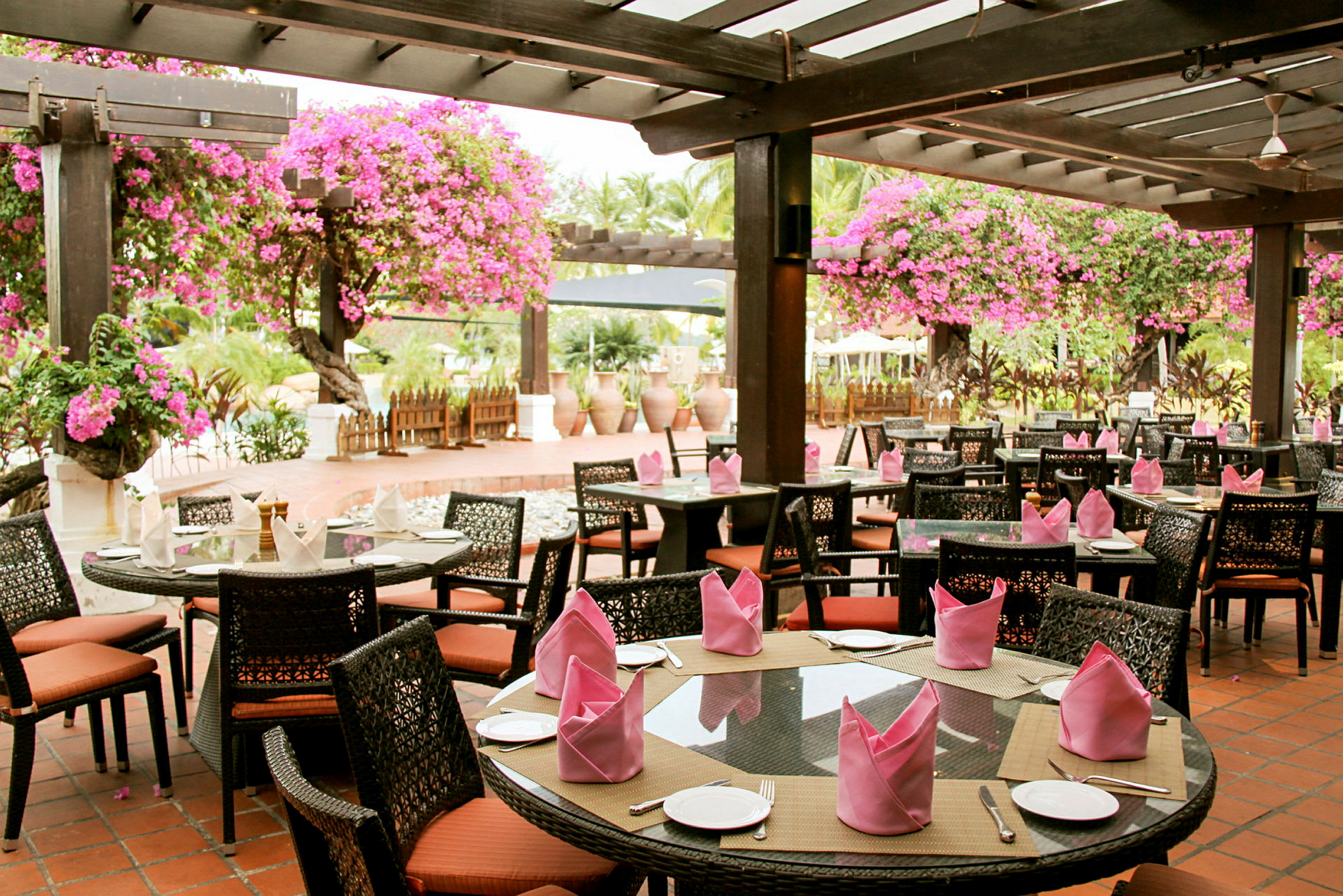 En utomhusrestaurang med bord och stolar, täckt av en pergola dekorerad med rosa blommor. (AI-genererad text)