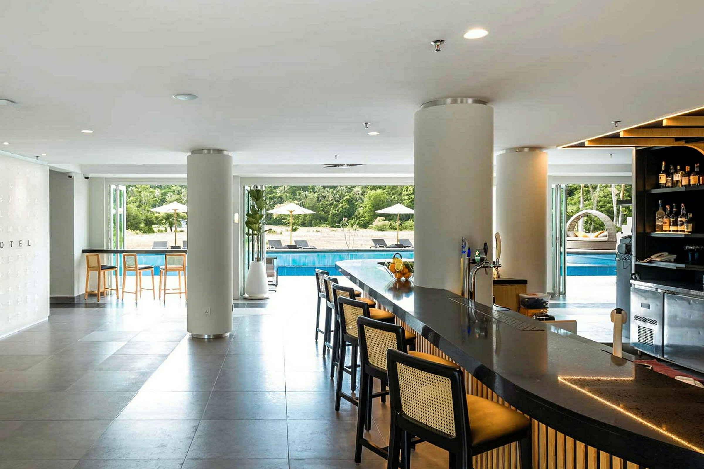 En hotellbar inomhus med utsikt över en utomhuspool omgiven av grönska. (AI-genererad text)
