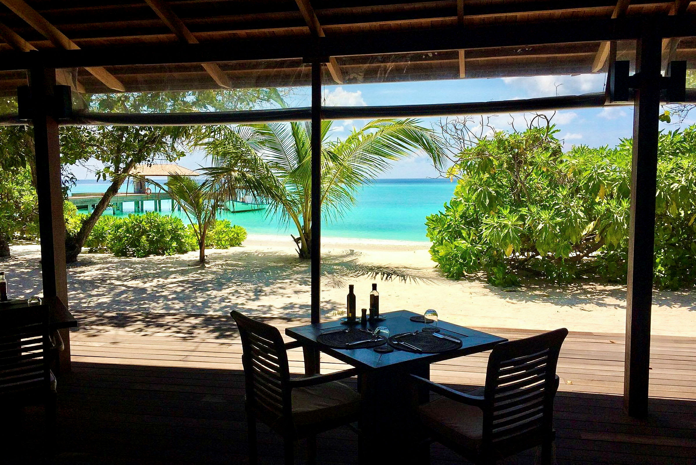 Utsikt från en restaurang mot en tropisk strand med turkost hav och grönskande vegetation. (AI-genererad text)