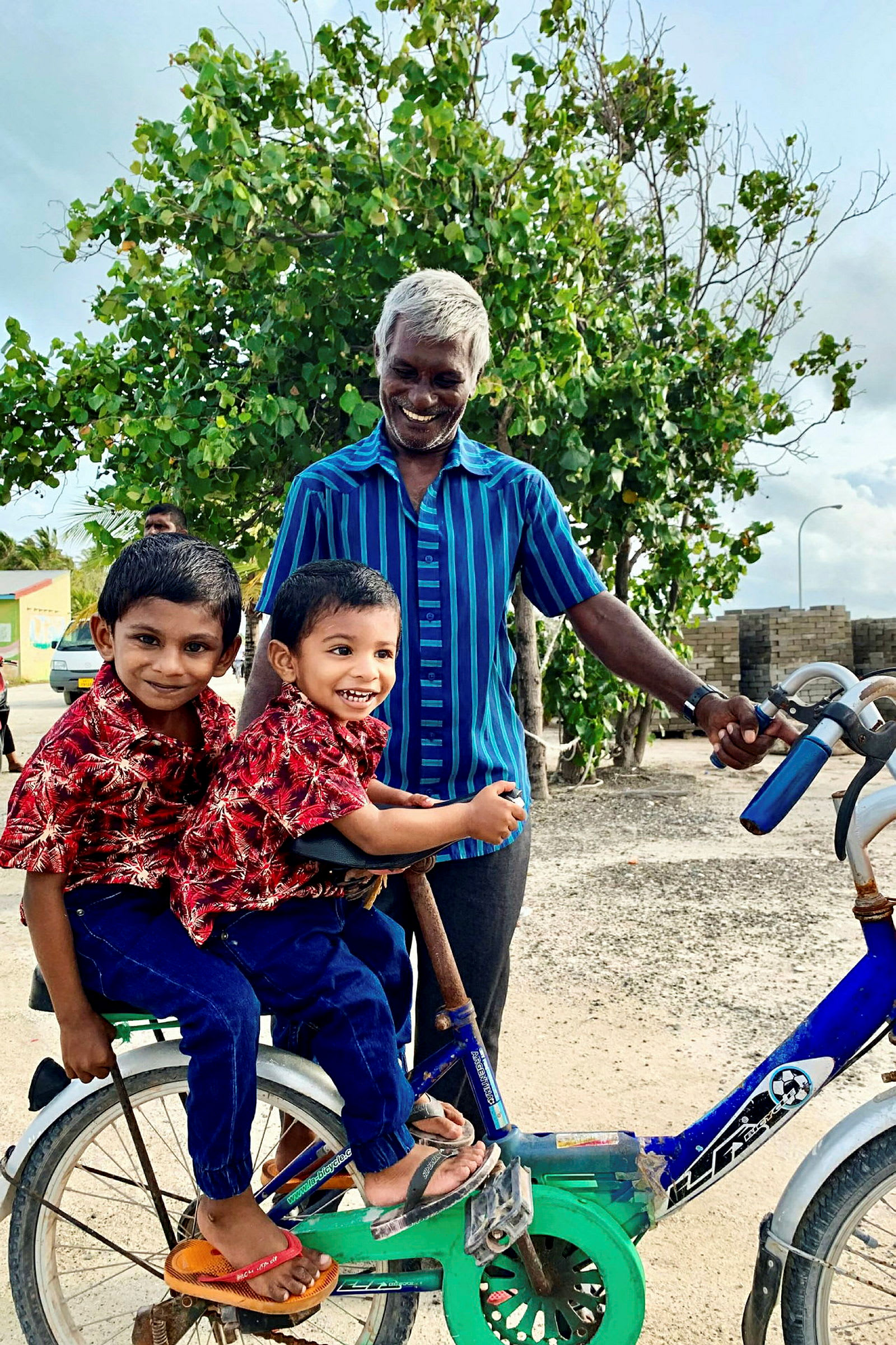 Två barn sitter på en cykel medan en vuxen håller i styret framför ett träd. (AI-genererad text)
