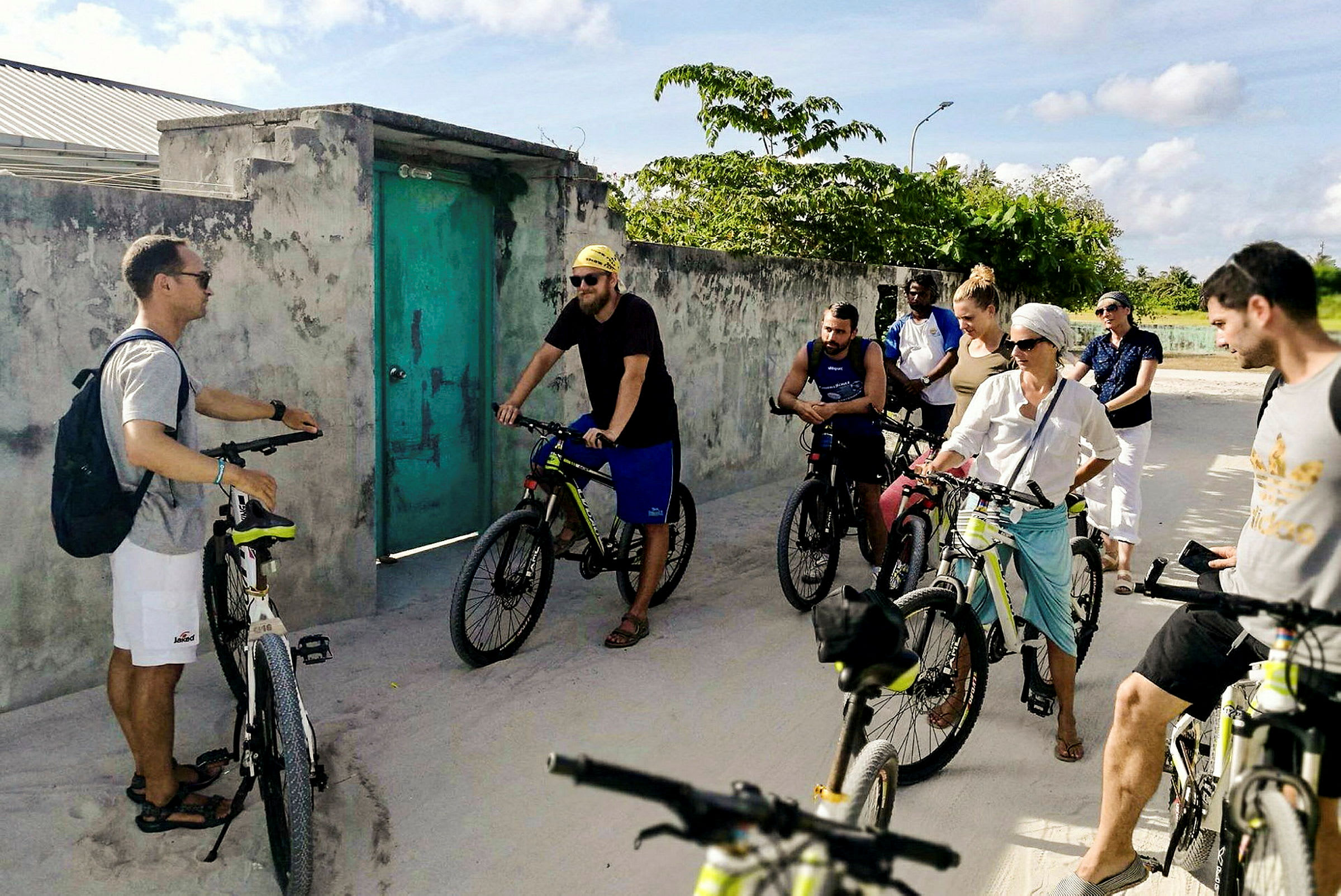 En grupp cyklister står vid en betongvägg med en grön dörr, omgivna av grönska. (AI-genererad text)