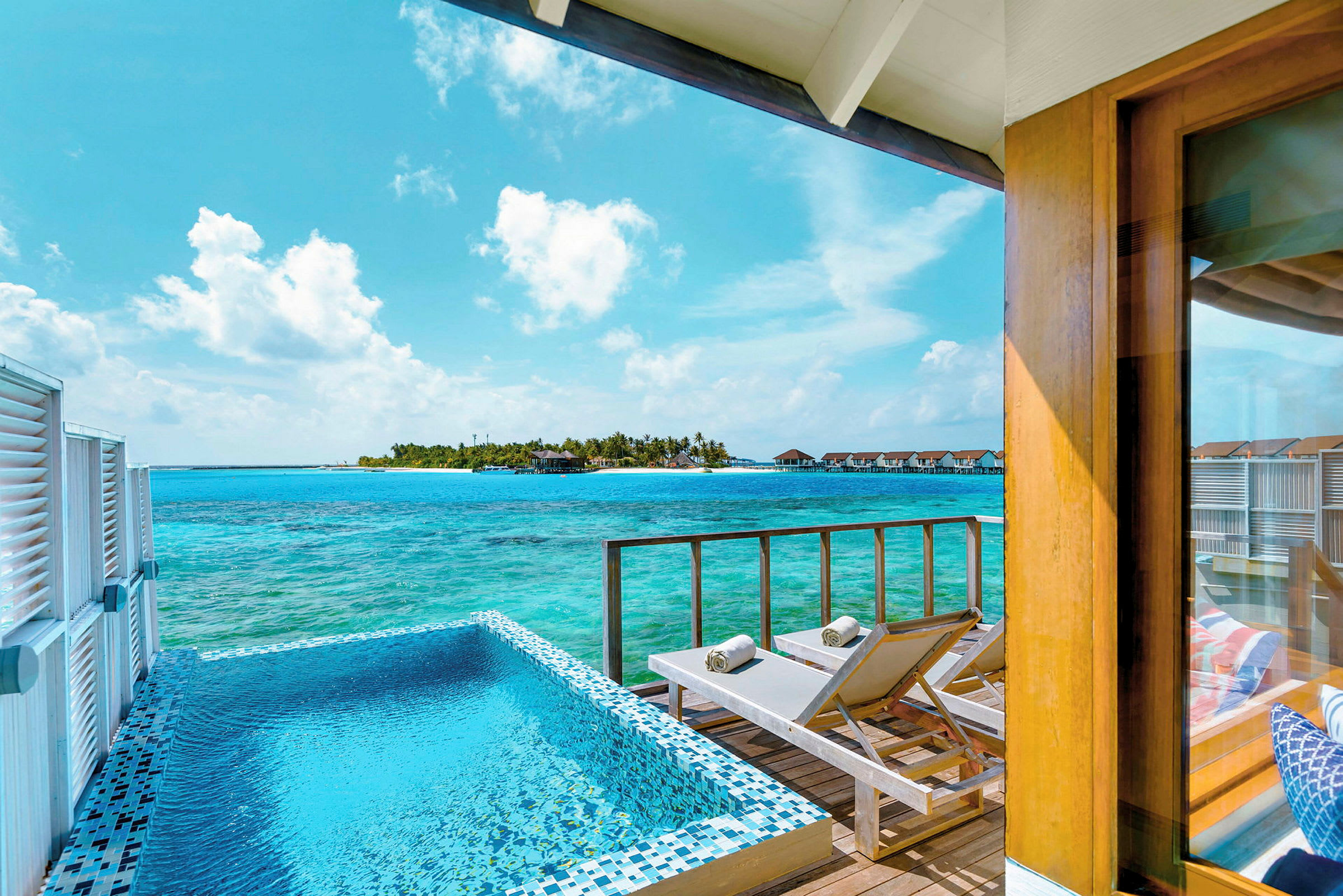 Utsikt från en terrass med pool och solstolar mot havet och en ö med bungalows. (AI-genererad text)