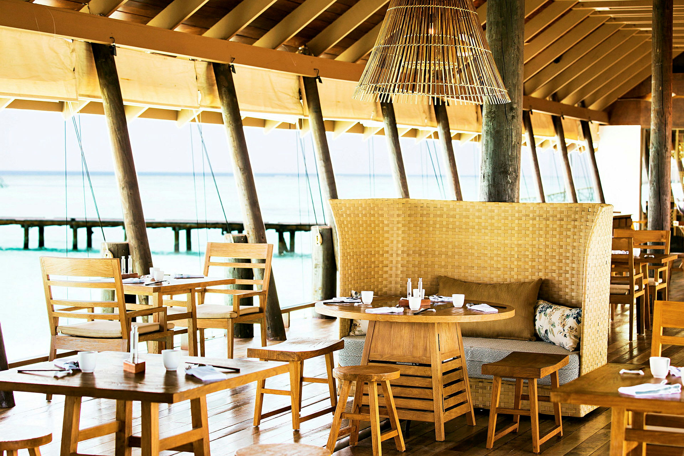 En restaurang med träbord och stolar samt en flätad bänk med kuddar, belägen vid havet. (AI-genererad text)