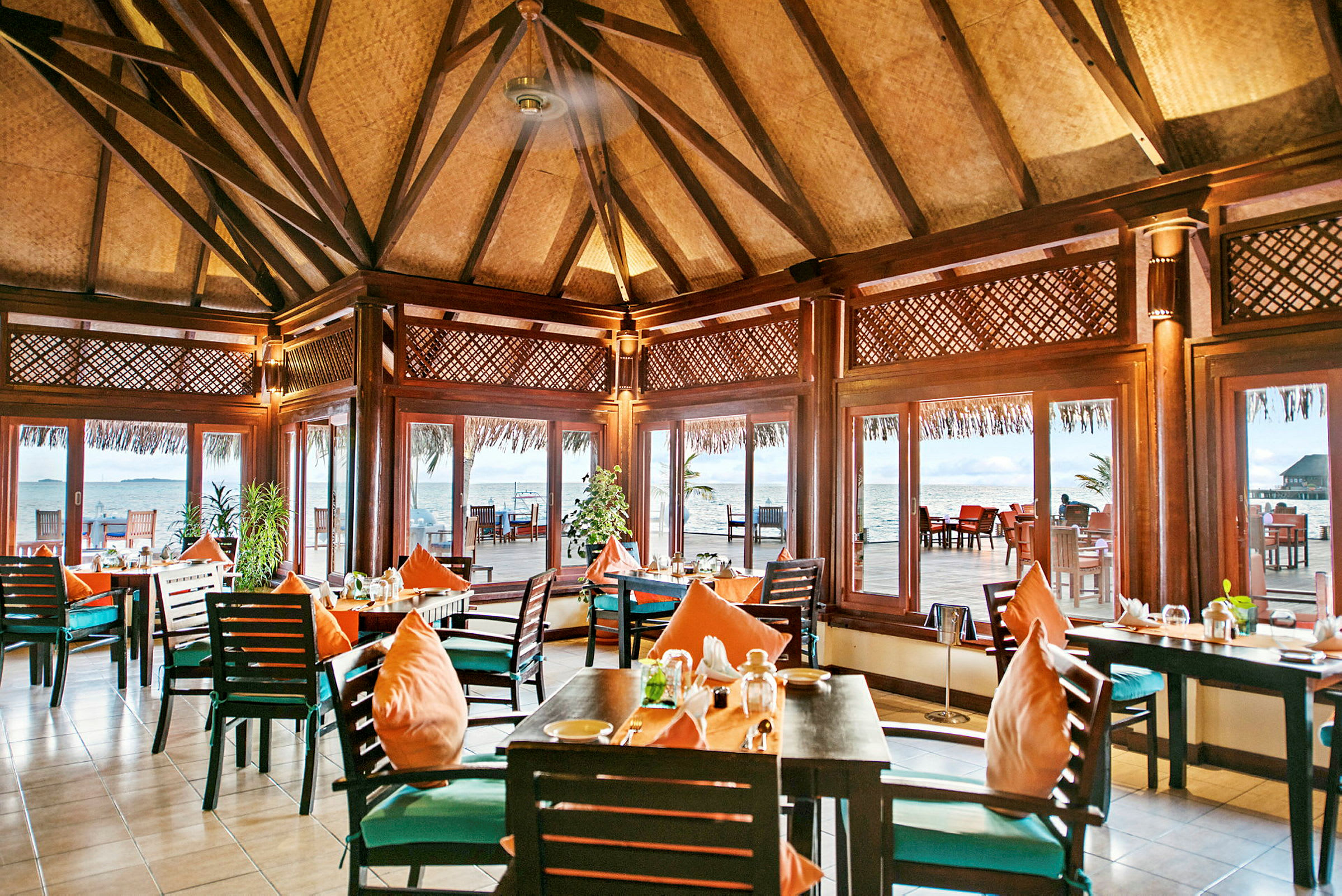 En restaurang med träinredning och bord med orange kuddar, omgiven av stora fönster med utsikt mot havet. (AI-genererad text)