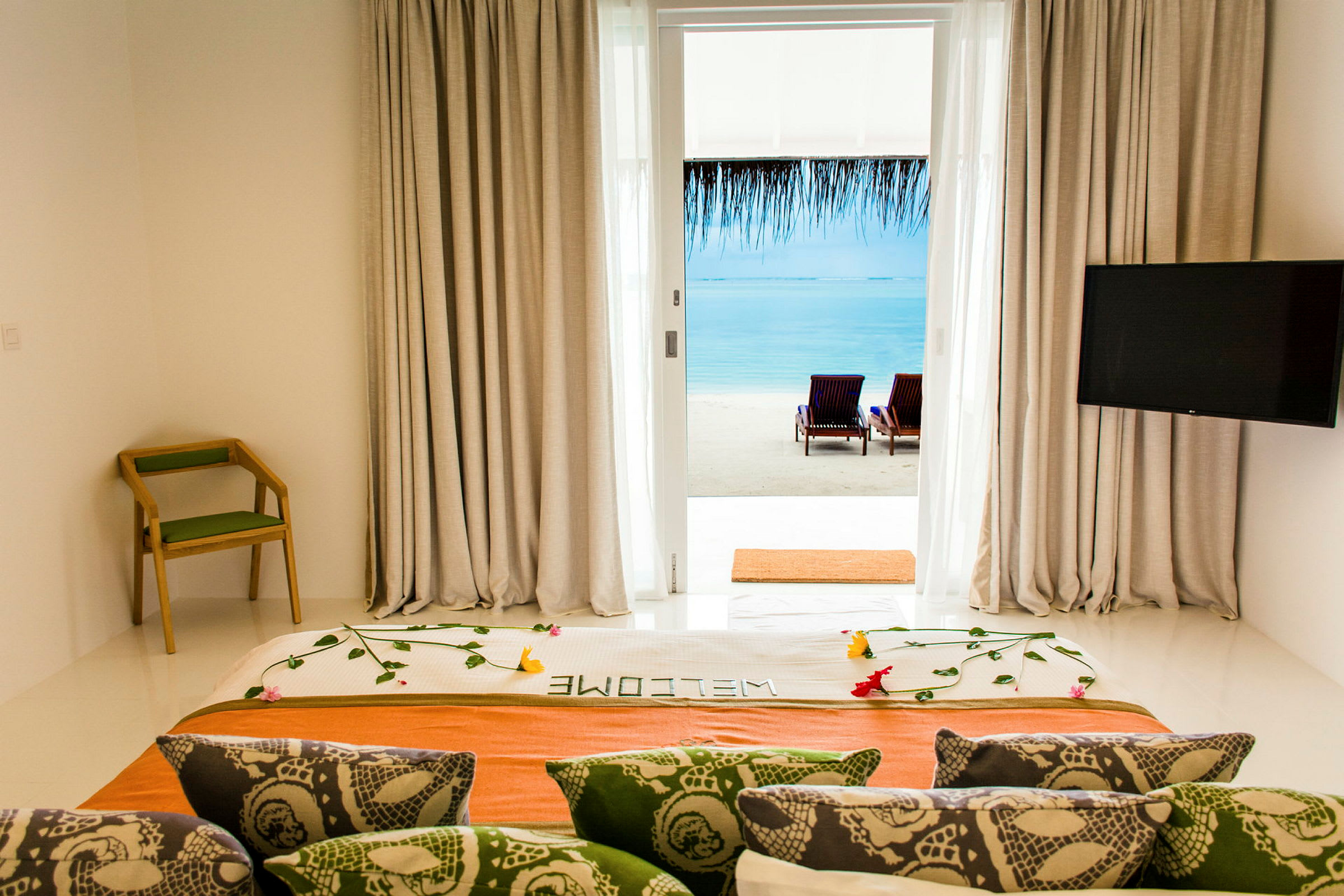 Utsikt från hotellrum med dekorerad säng, ryggstödsstol, och TV, mot strand och hav genom glasdörr. (AI-genererad text)