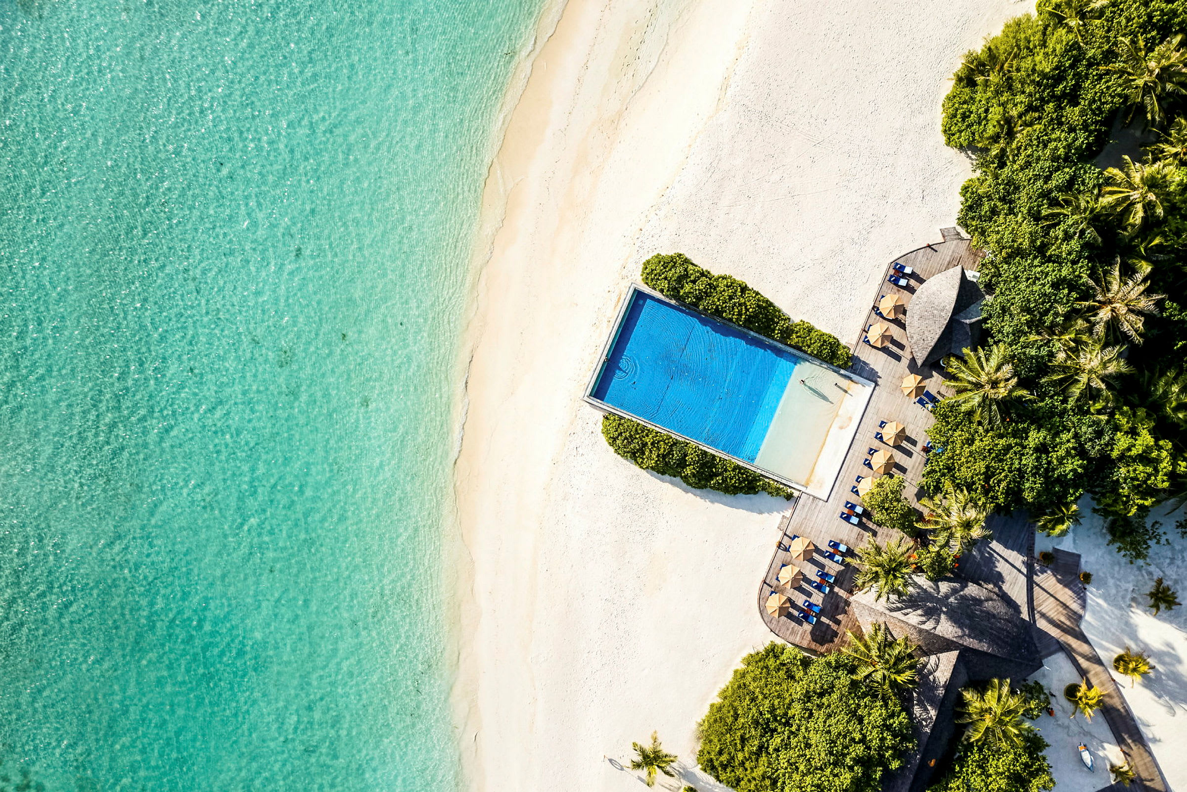 Flygbild av en strand, en pool omgiven av grönska och solstolar med parasoller. (AI-genererad text)