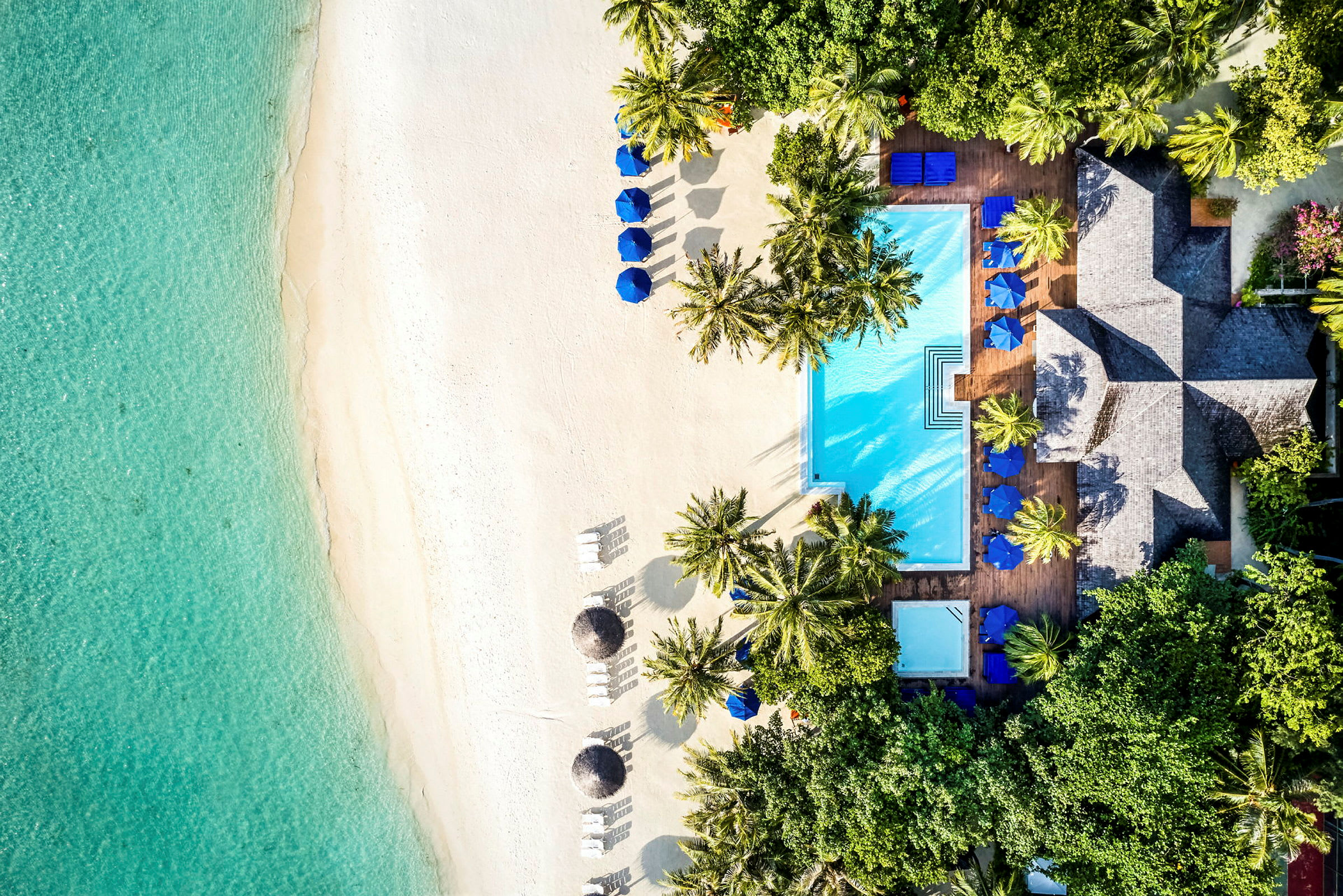 Flygbild av en strand med vit sand, turkosfärgat vatten, parasoller och en pool omgiven av palmträd. (AI-genererad text)