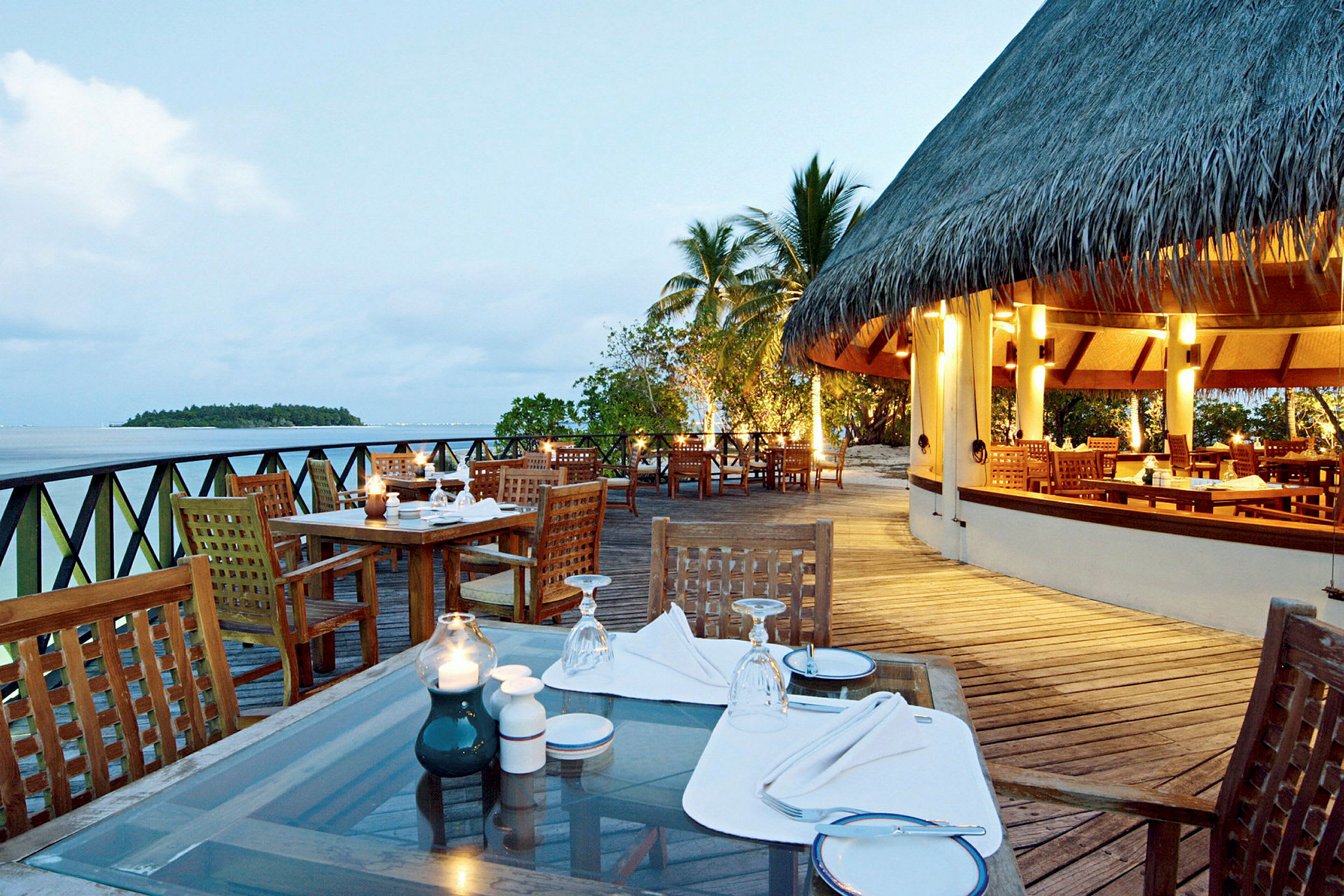 En öppen restaurang vid stranden med trämöbler och dukade bord, ö och hav syns i bakgrunden. (AI-genererad text)