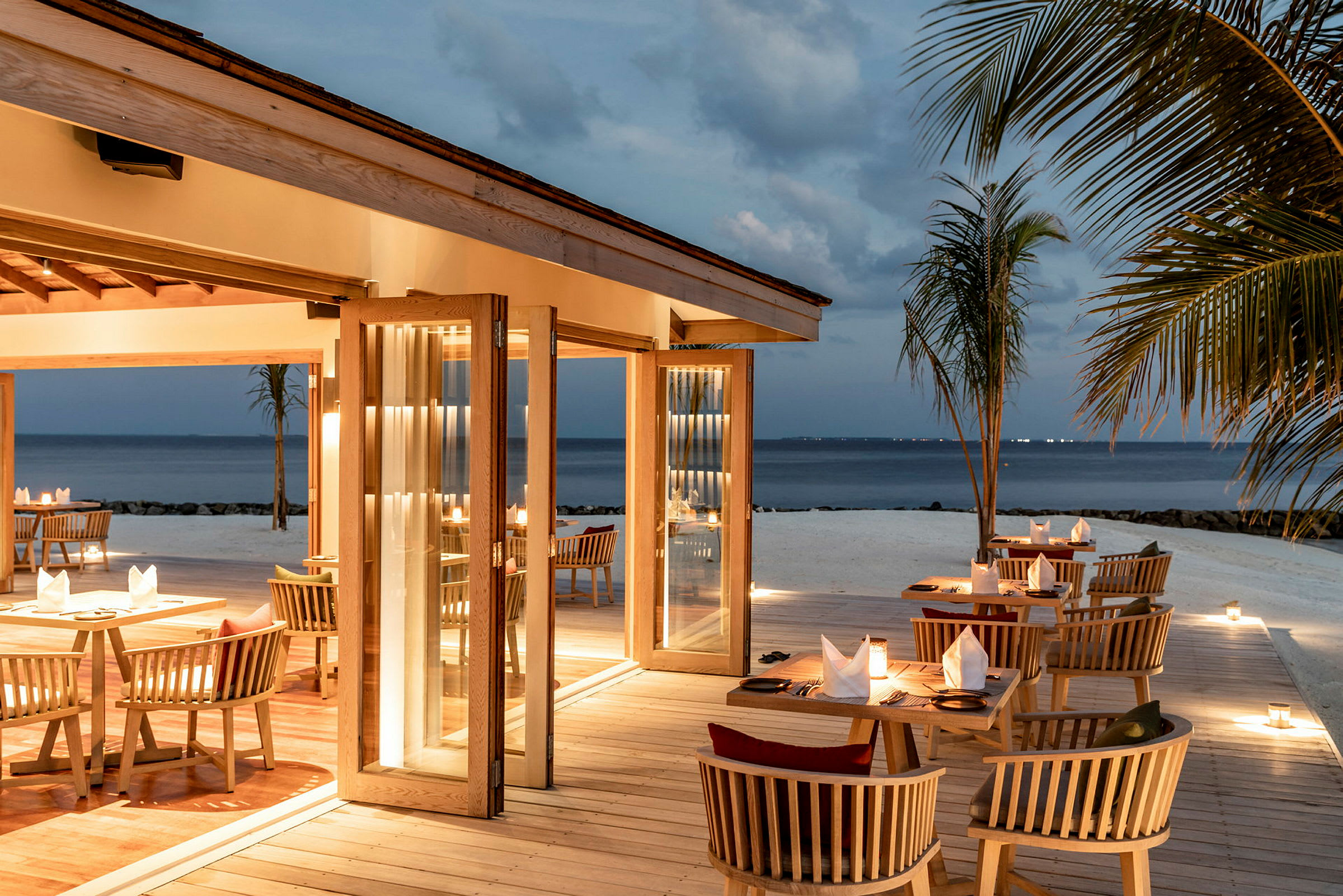 Utomhusrestaurang med bord och stolar vid stranden under skymning. (AI-genererad text)