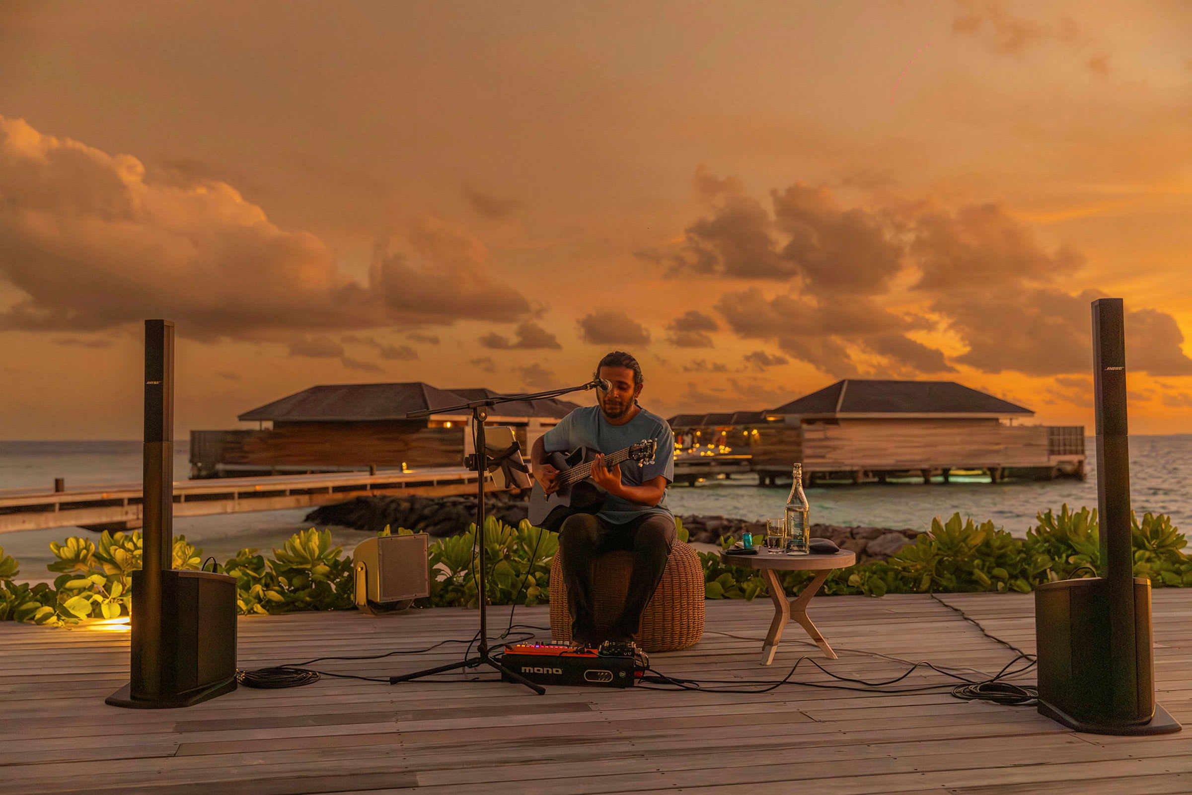 En person spelar gitarr medan solen går ned vid en strand med vattenbungalows. (AI-genererad text)