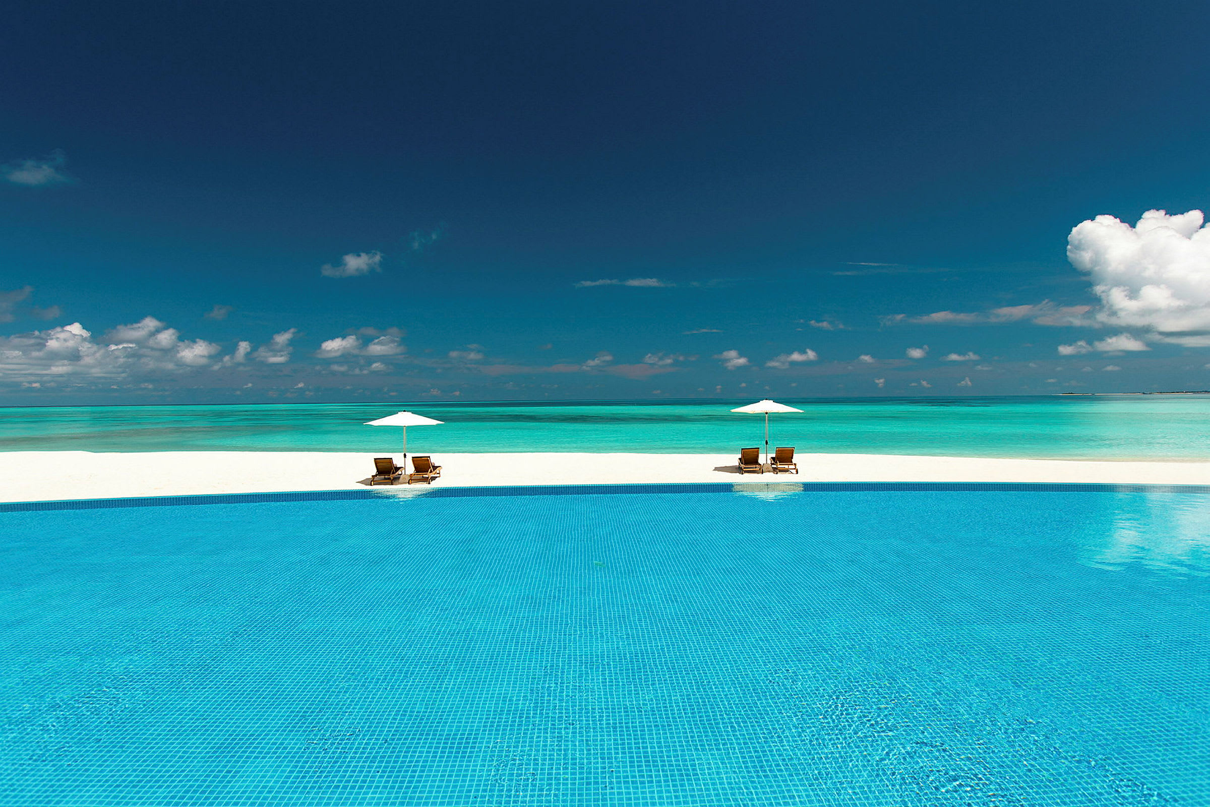 En infinitypool framför en paradisstrand med två solstolar och parasoller. (AI-genererad text)