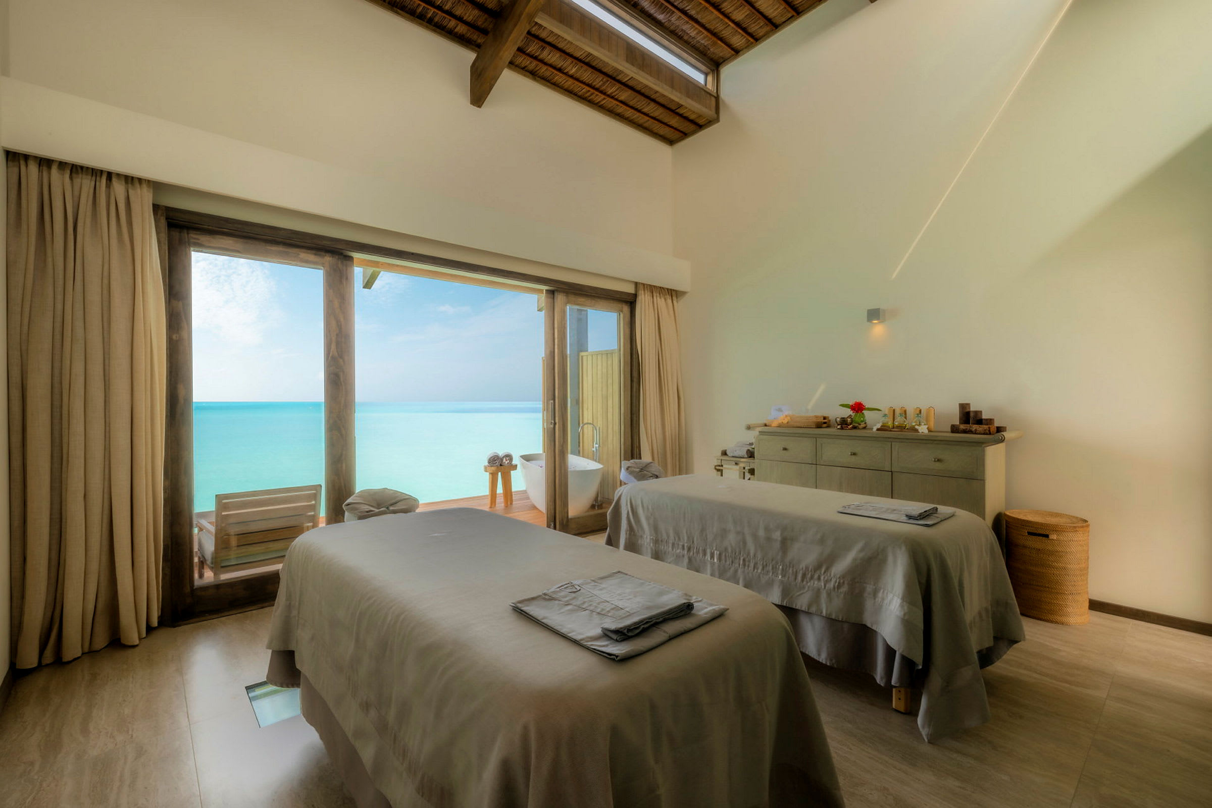 Ett ljust behandlingsrum med två massagebord och stora fönster som vetter ut mot en veranda och havet. (AI-genererad text)