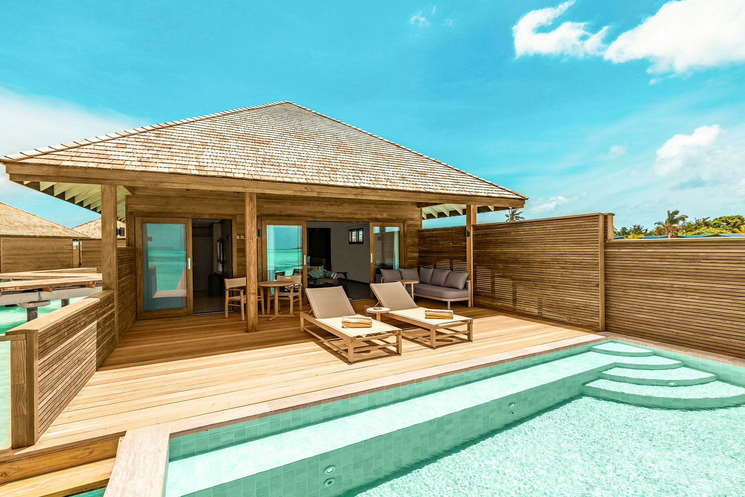 En terrass med två solstolar, bord och en privat pool framför en bungalow i trä. Soligt väder och klart blå himmel. (AI-genererad text)