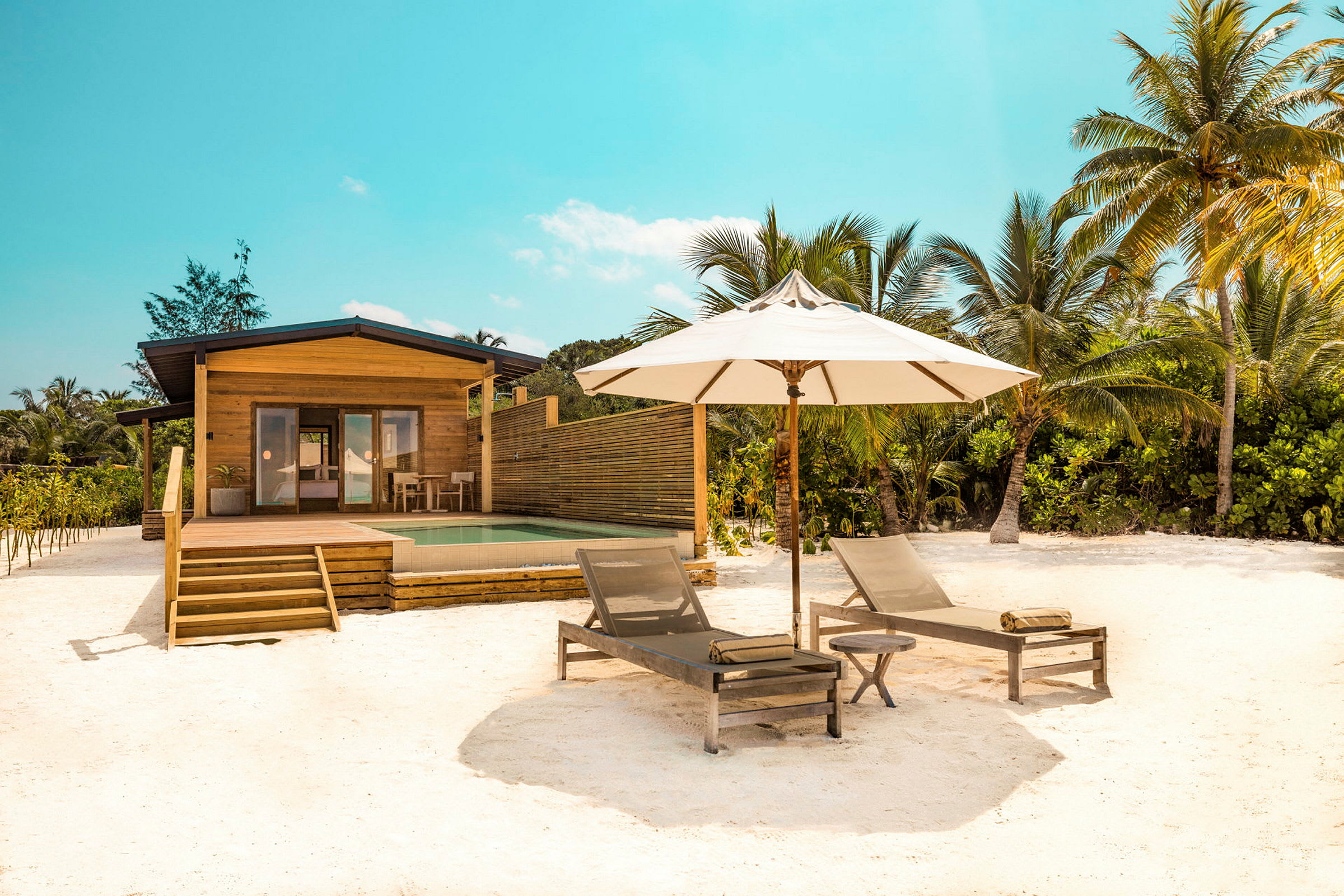 Ett sommarhus framför en pool med två solstolar och ett parasoll på en sandstrand. (AI-genererad text)