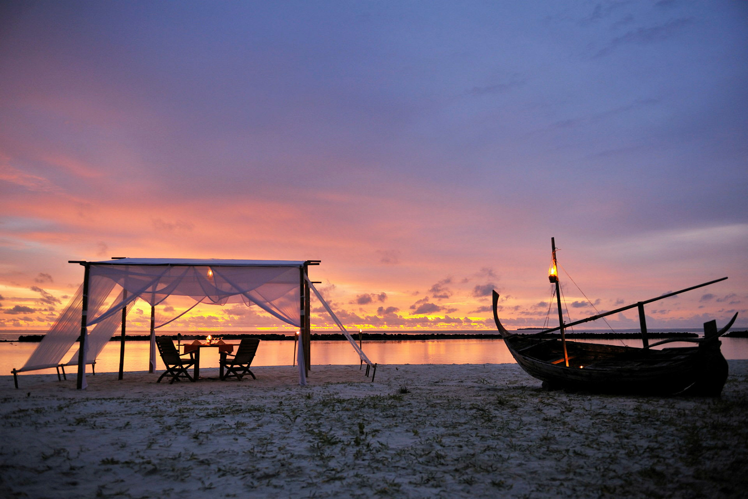 En strand med en liten baldakin och stolar vid solnedgången, med en träbåt och lyktor. (AI-genererad text)