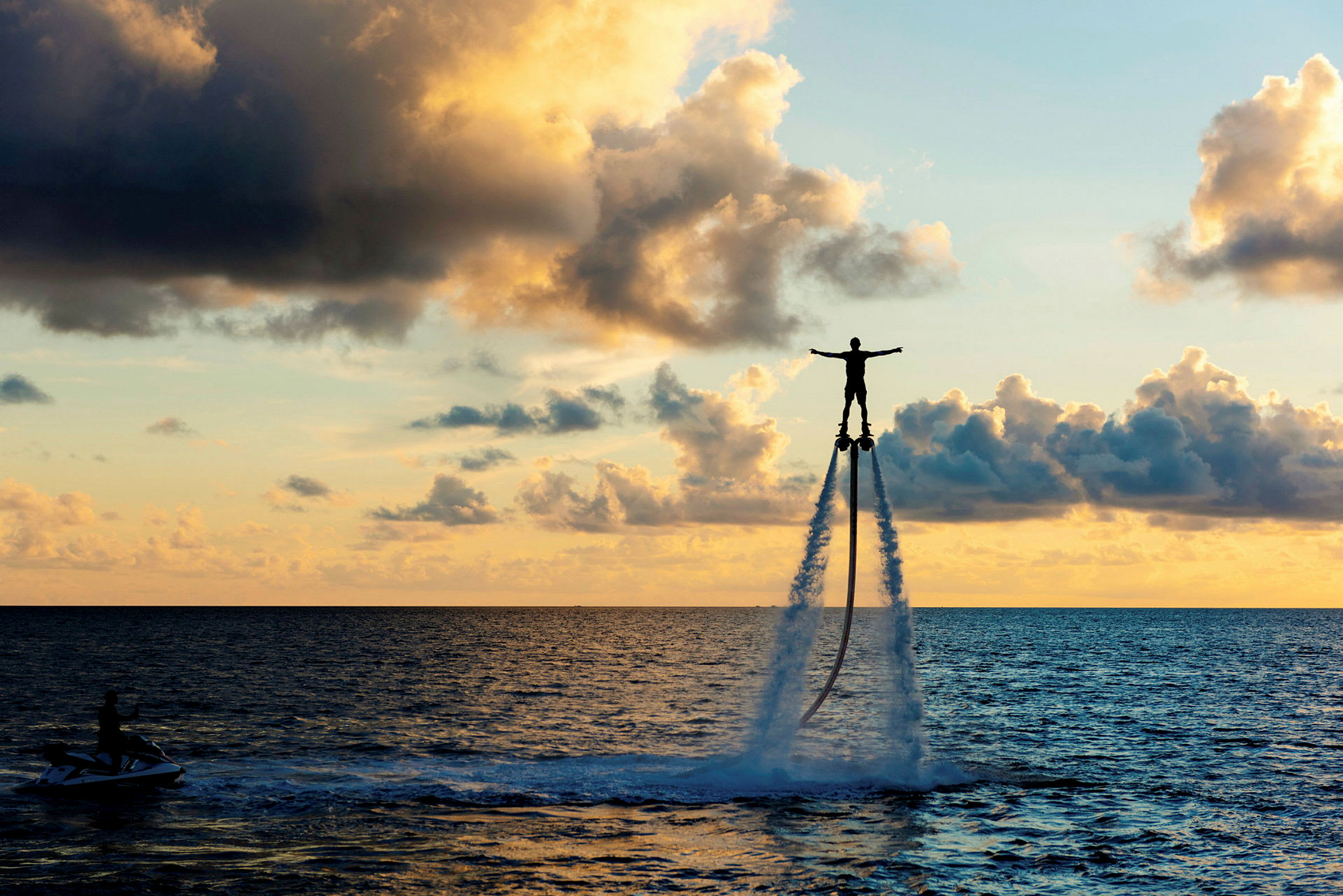 En person flyger ovanför havet med hjälp av en vattenjetpack medan solen går ned. (AI-genererad text)