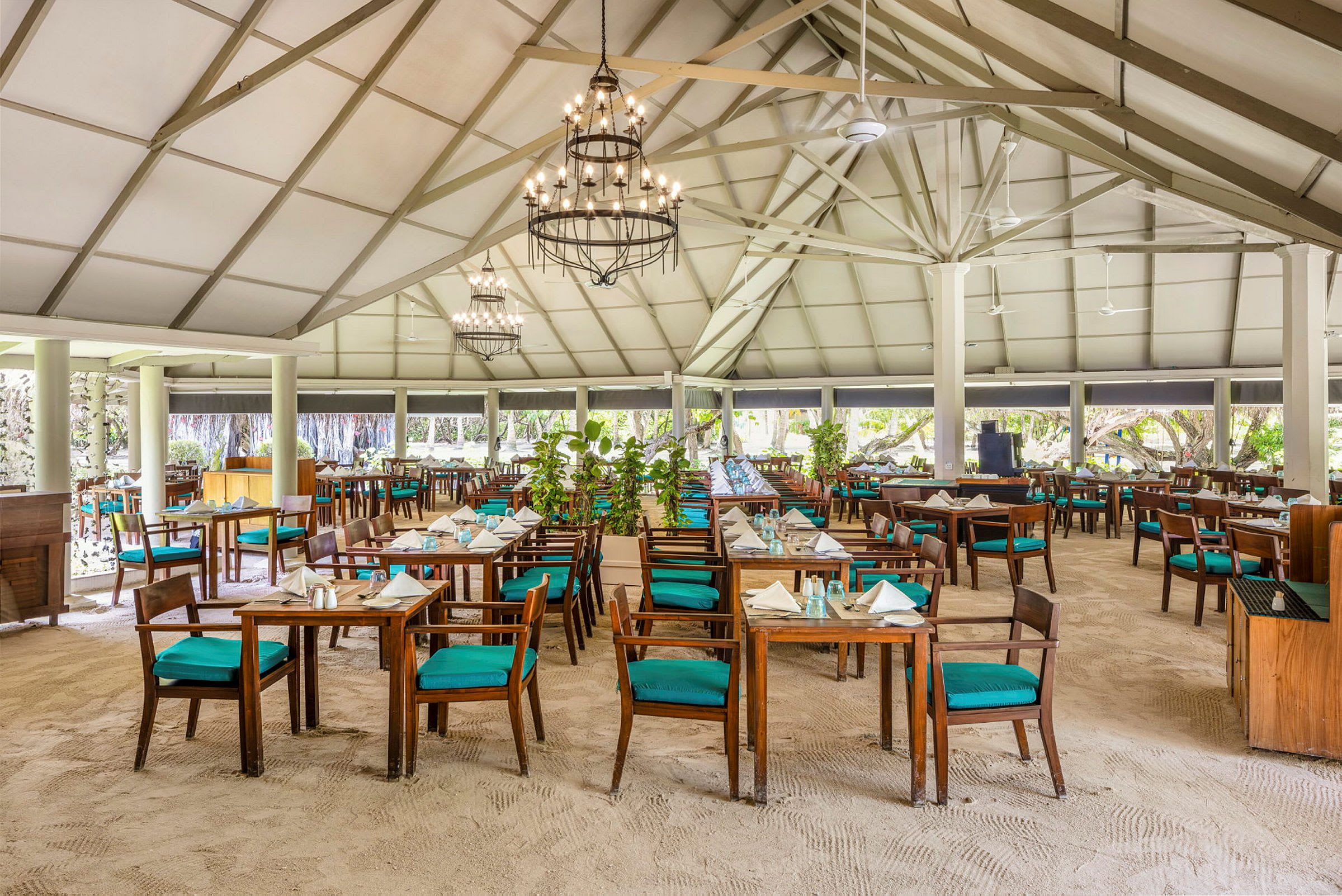 En rymlig restaurang med sandgolv och bord med trästolar klädda med turkosa dynor, under ett högt tak med takbjälkar. (AI-genererad text)