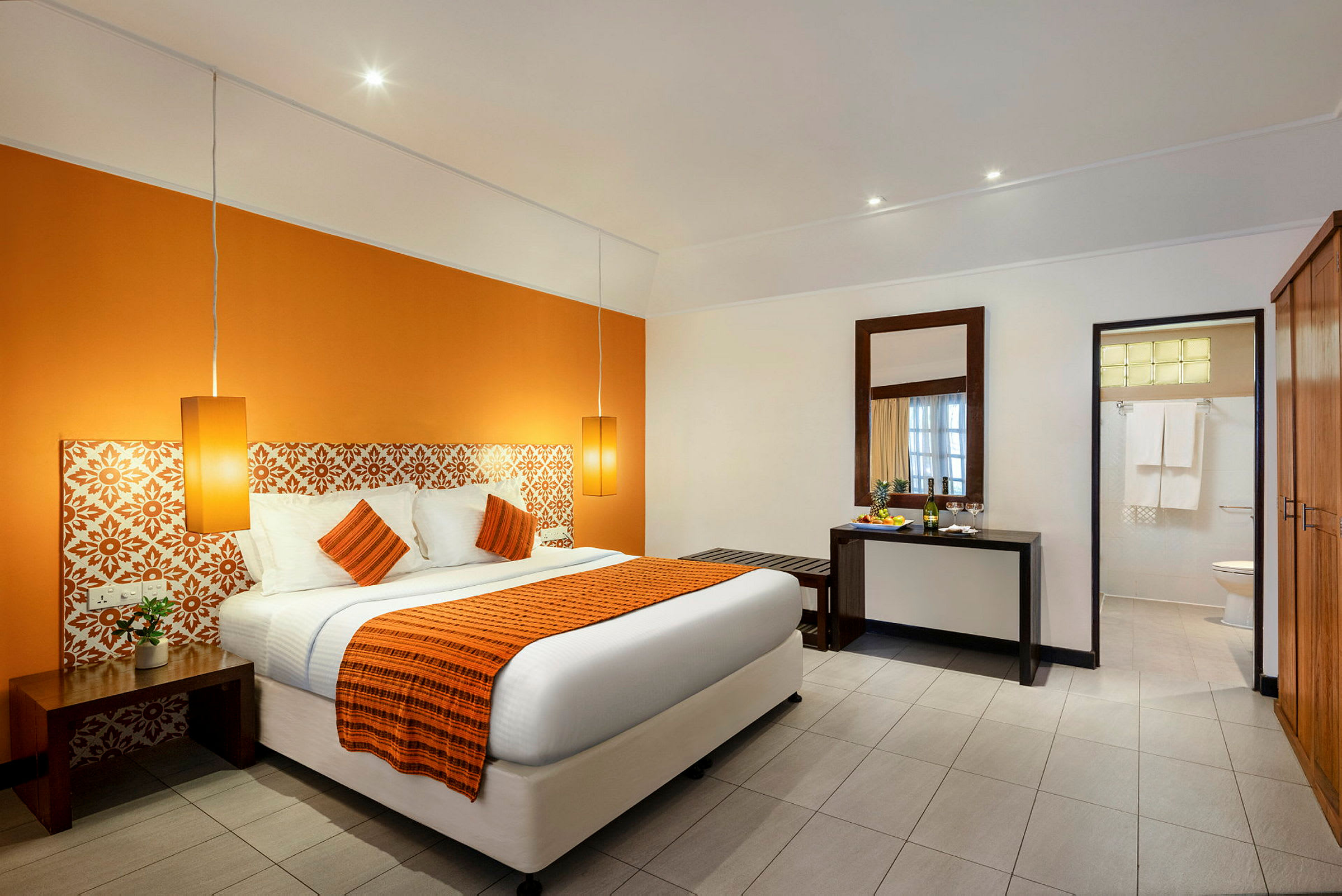 Ett hotellrum med en dubbelsäng med orange överkast och prydnadskuddar, två hängande lampor, ett bord med spegel och en dörr till ett badrum. (AI-genererad text)