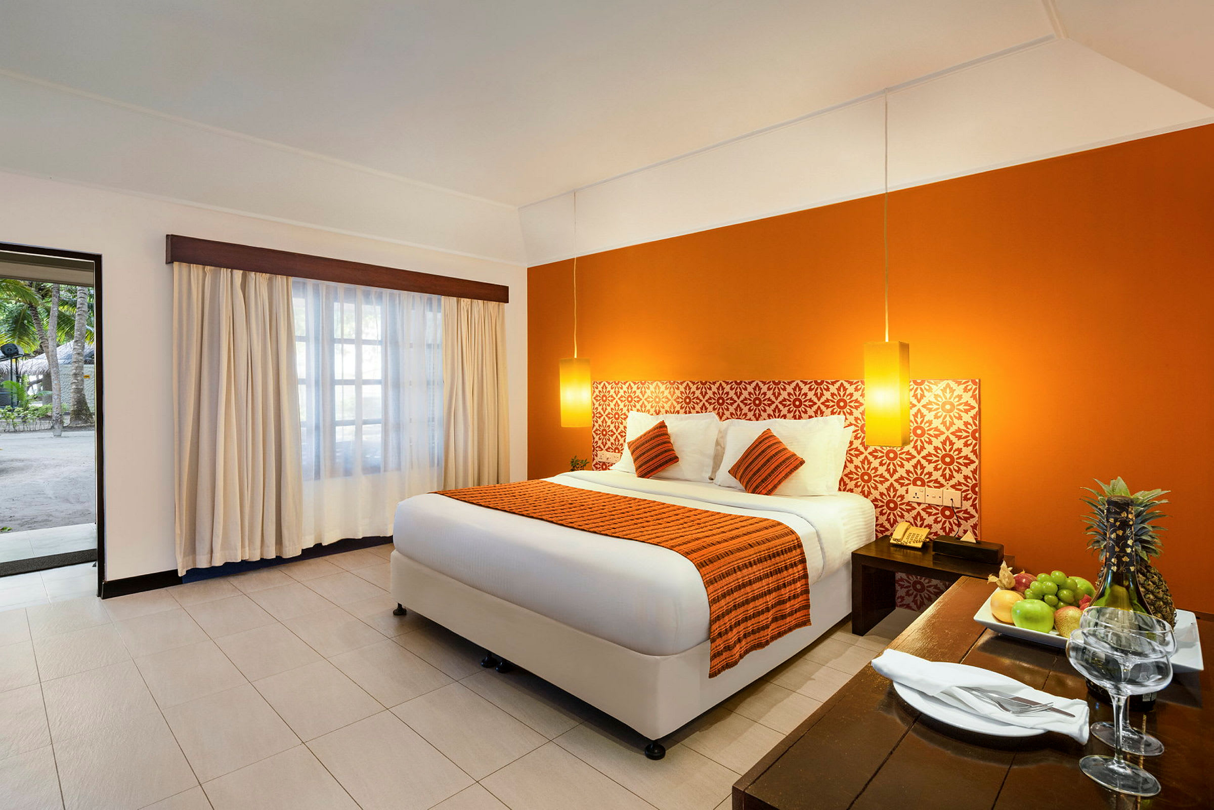 Ett hotellrum med en dubbelsäng med orange detaljer, ett bord med frukt och glas, och ett fönster som vetter mot en utomhusmiljö. (AI-genererad text)