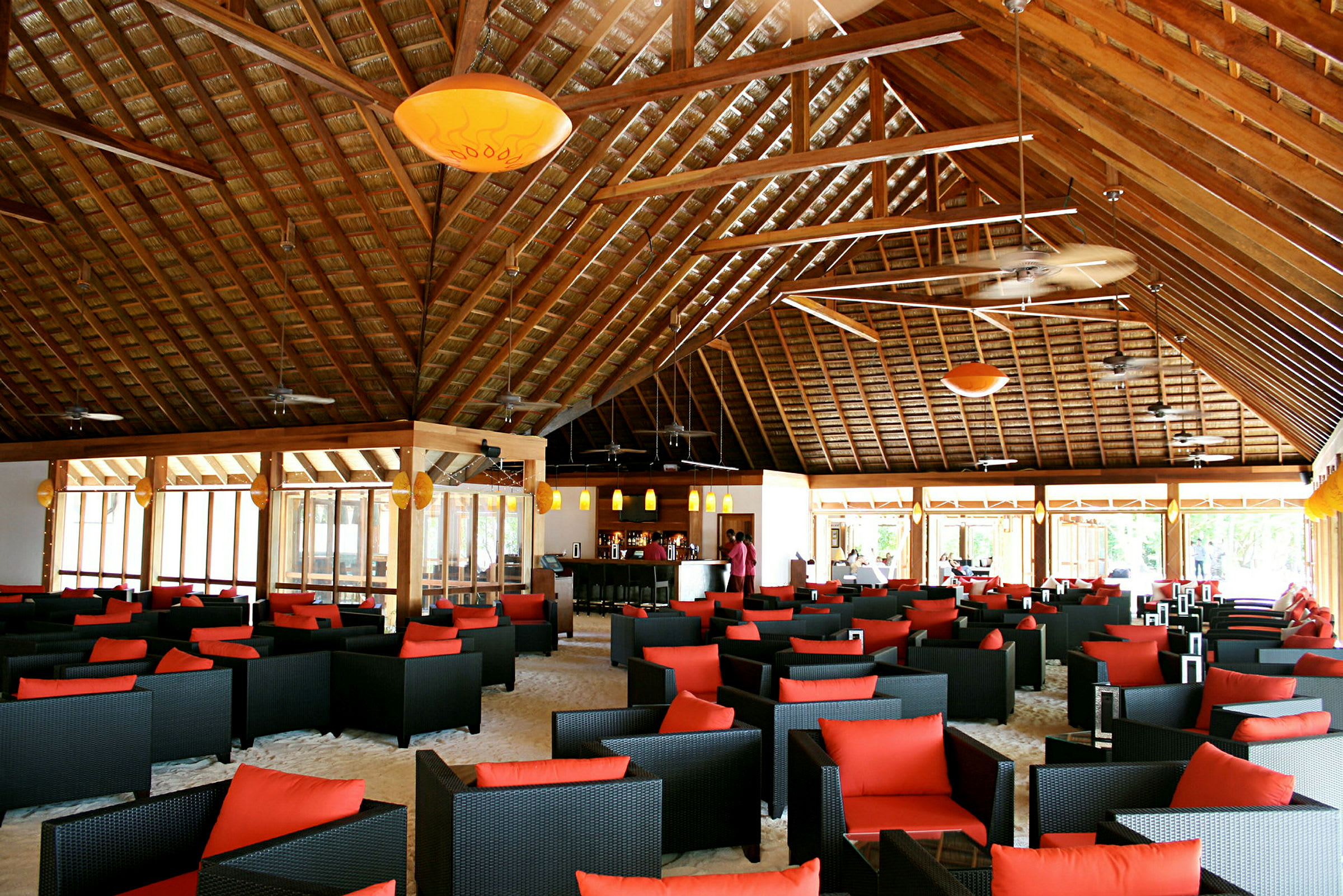 En interiör av en rymlig restaurang eller lounge med svarta flätade stolar med röda kuddar, bord placerade på sandgolv under ett högt träbjälktak. (AI-genererad text)