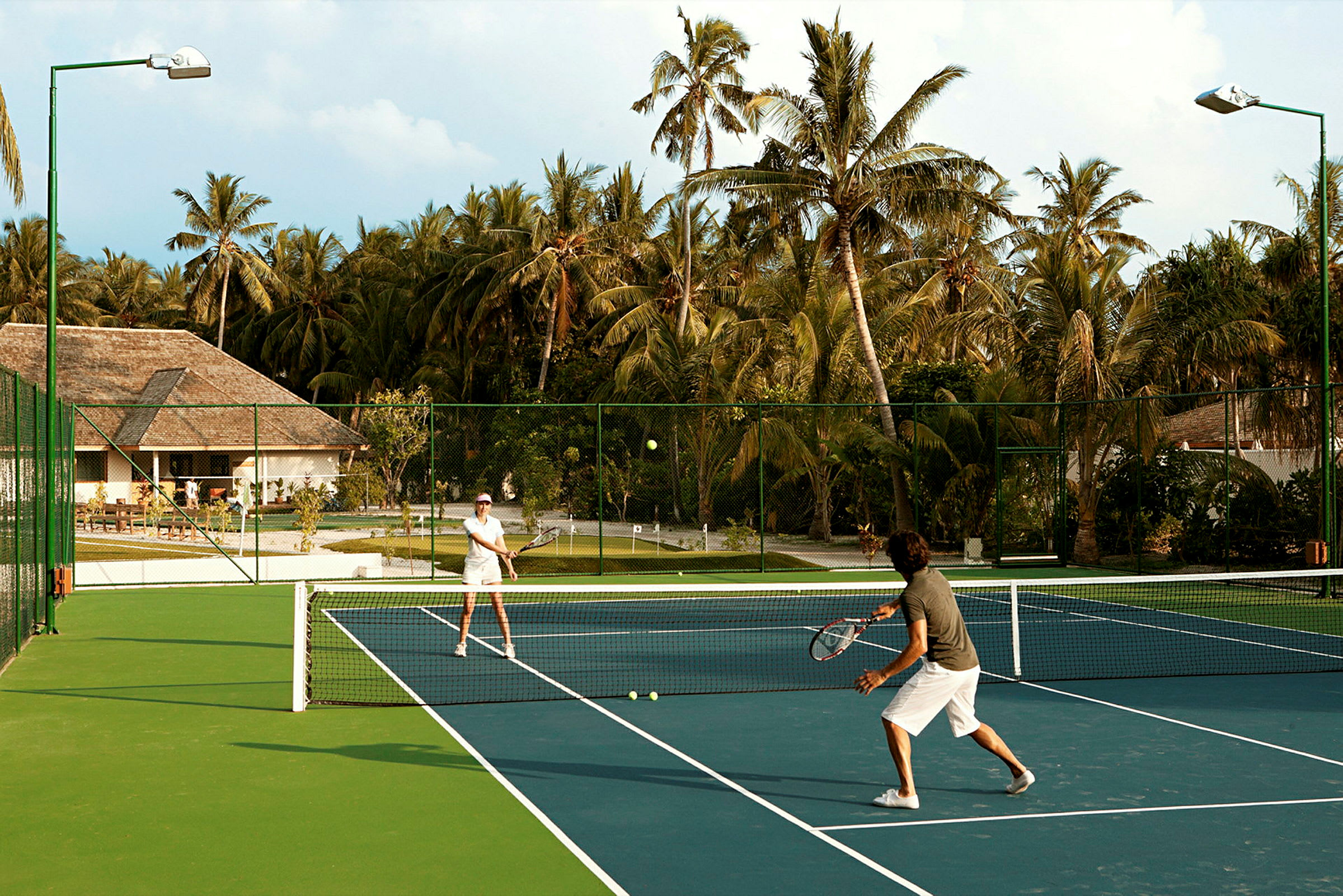Två personer spelar tennis på en utomhusbana omgiven av palmer. (AI-genererad text)
