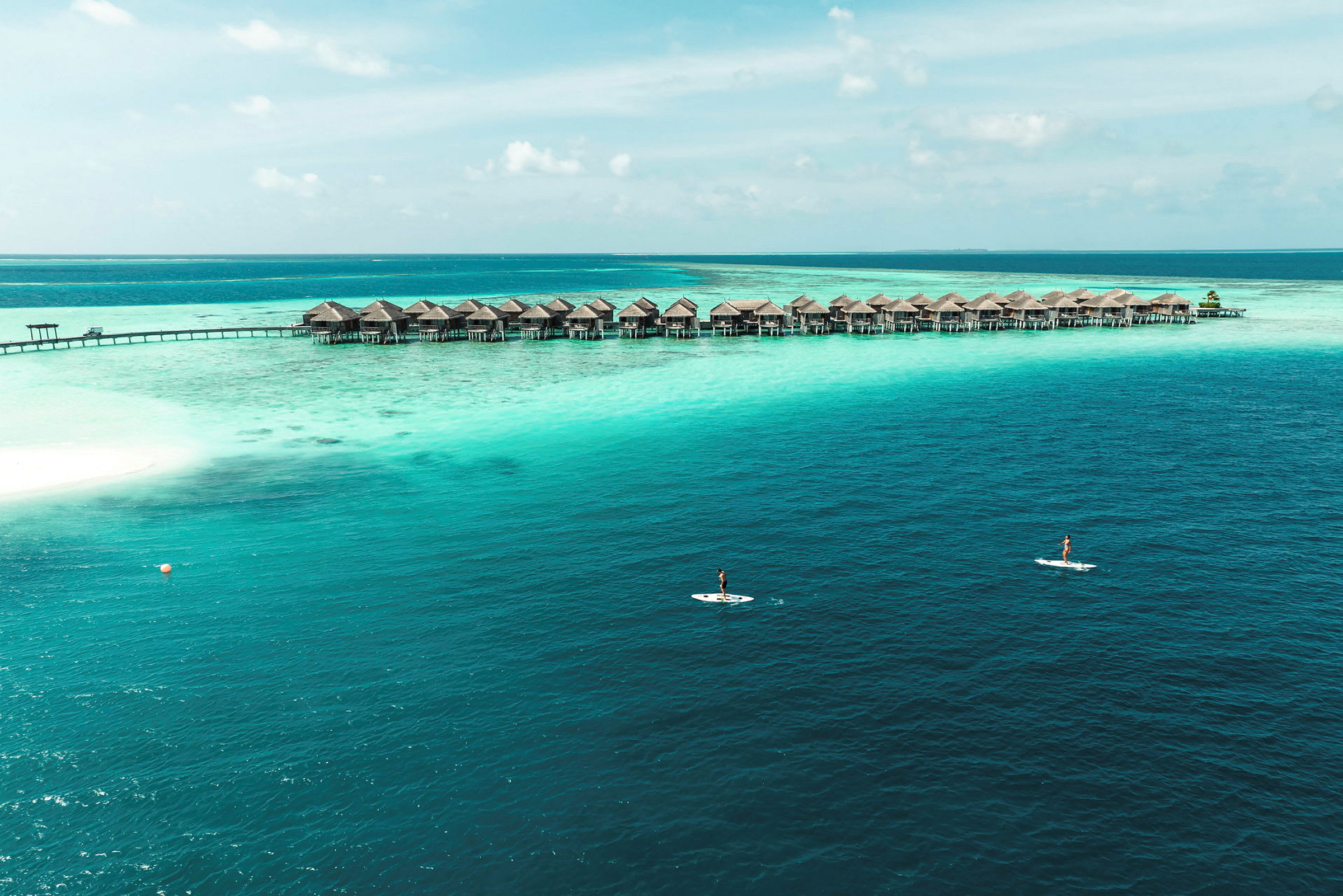 Övervattenbungalower på pålar vid ett turkost hav med en person på en paddleboard i förgrunden. (AI-genererad text)