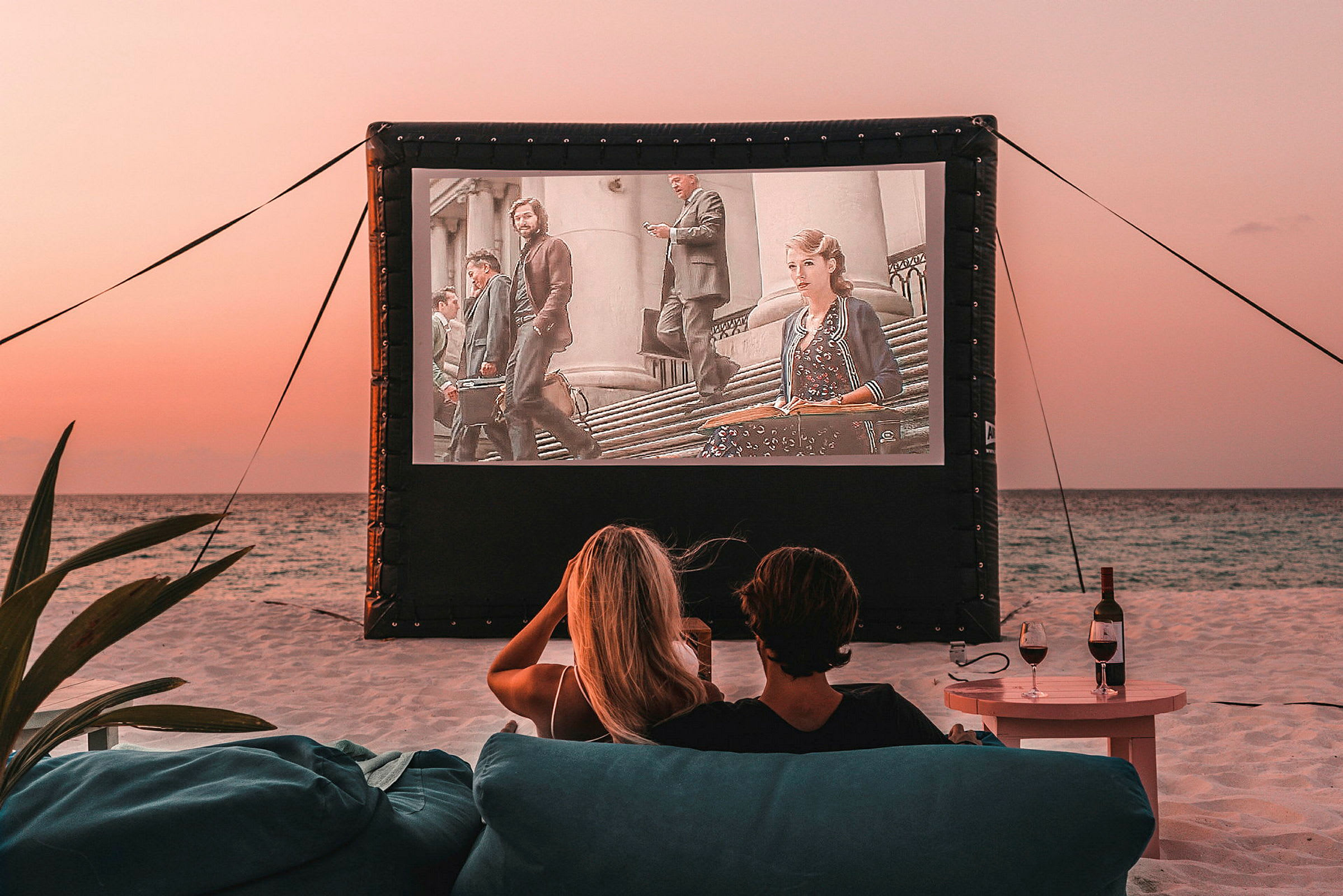 Två personer sitter på stranden och tittar på en film som visas på en utomhusduka. (AI-genererad text)