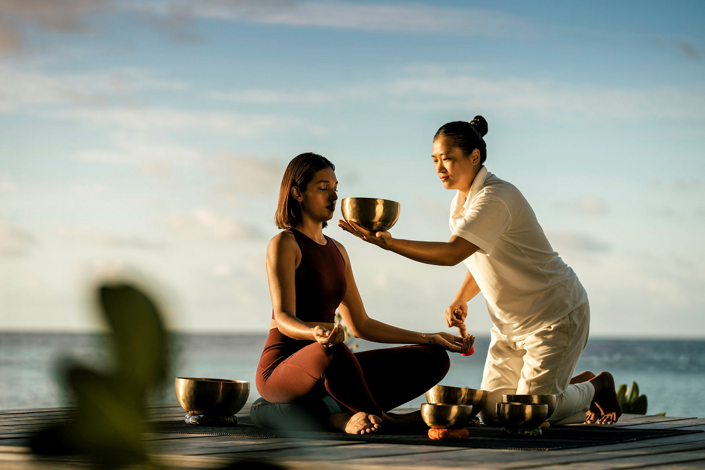 Två personer är del av ett meditationstillfälle nära vatten. En håller i en klangskål och en sitter med korslagda ben. (AI-genererad text)